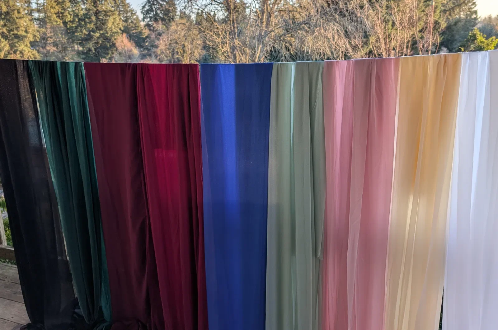 Floor-length table linens hanging side by side in black, dark green, burgundy, navy, sage, mauve pink, champagne and white.