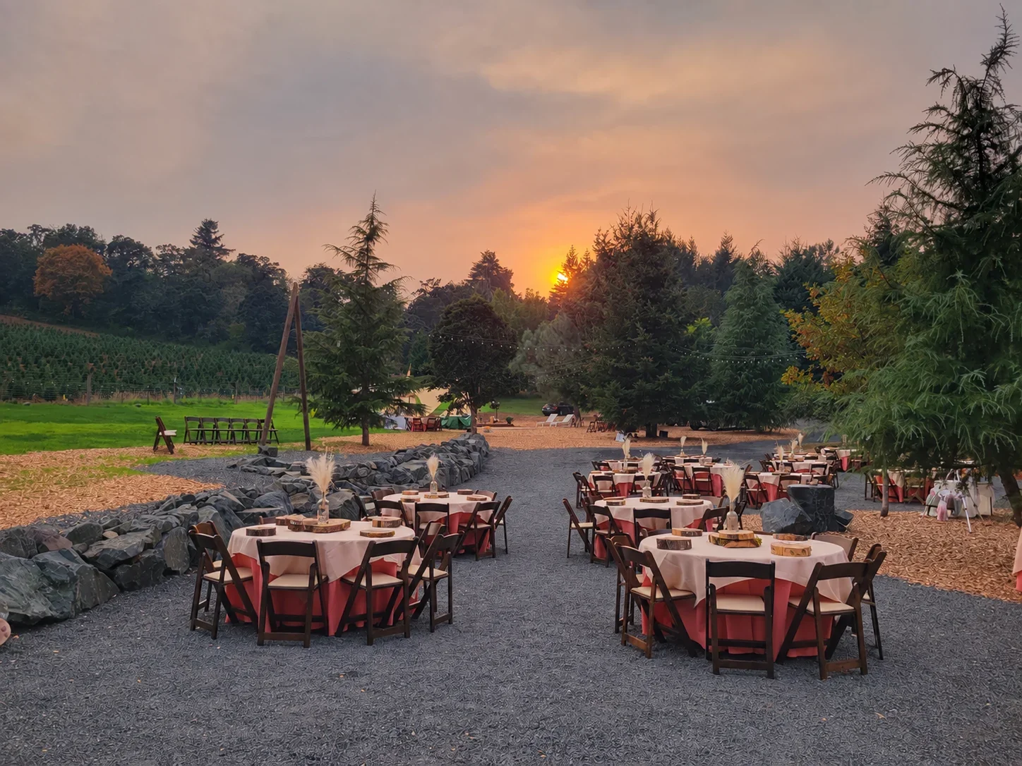 Round tables with coral floor-length linens, champagne overlays and wood slice centerpieces, surrounded by dark wood folding chairs at an outdoor event at sunset.