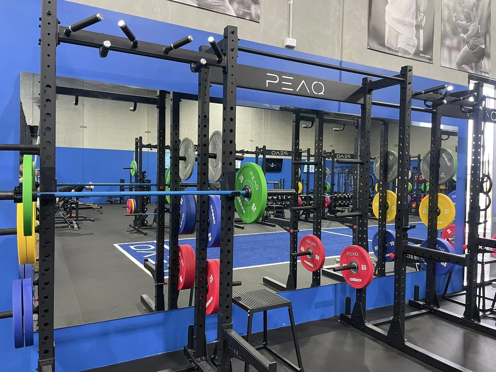 A gym with black metal weight racks holding colorful weight plates, including yellow, green, blue, and red. There is a blue workout platform and posters of athletes on the gray wall behind. The gym has a modern, organized look.