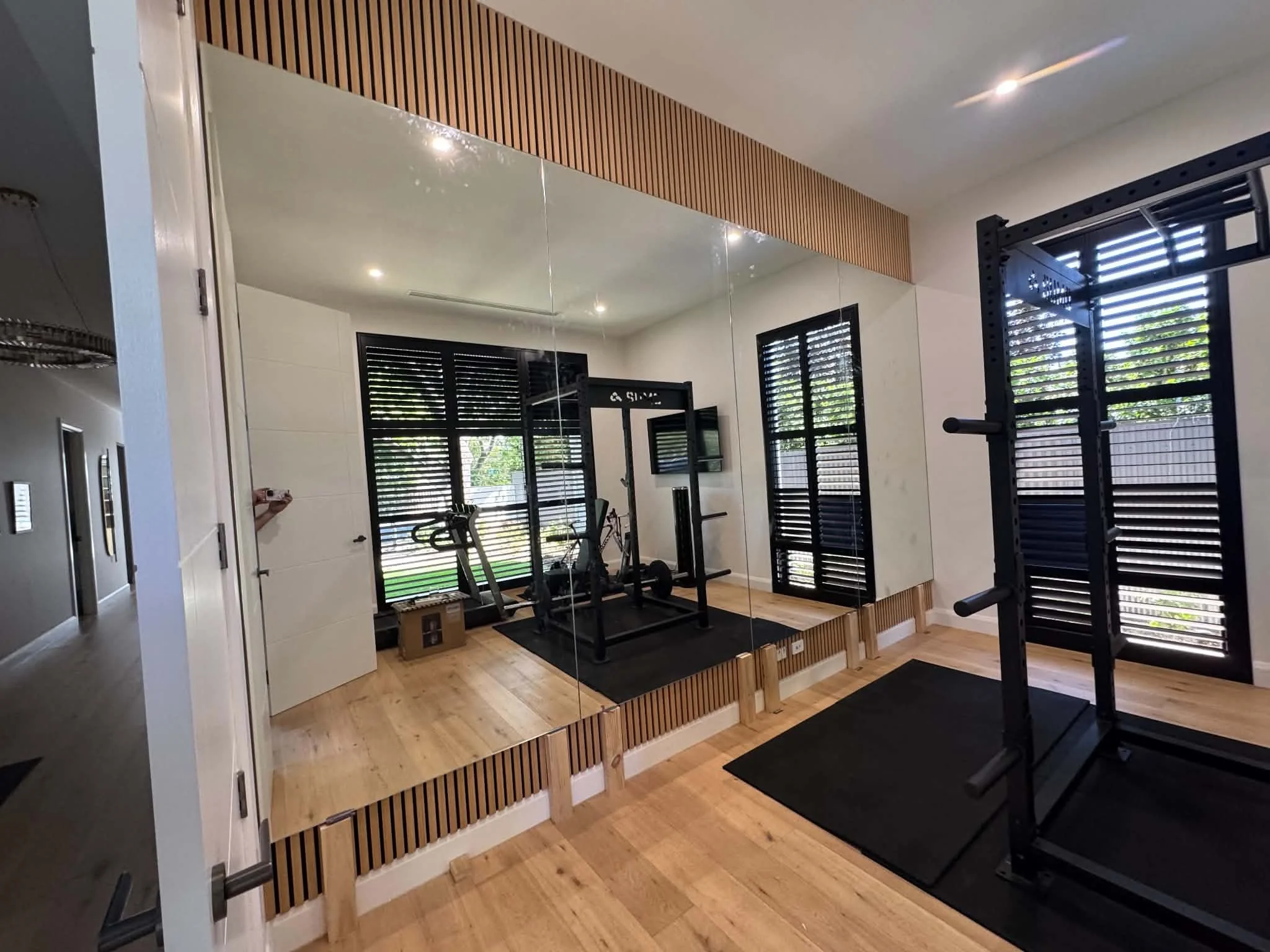 A home gym room with a large mirror on the wall, black workout equipment including a squat rack, and a window with black shutters. The room has wooden flooring and decorative wooden slats on the wall.