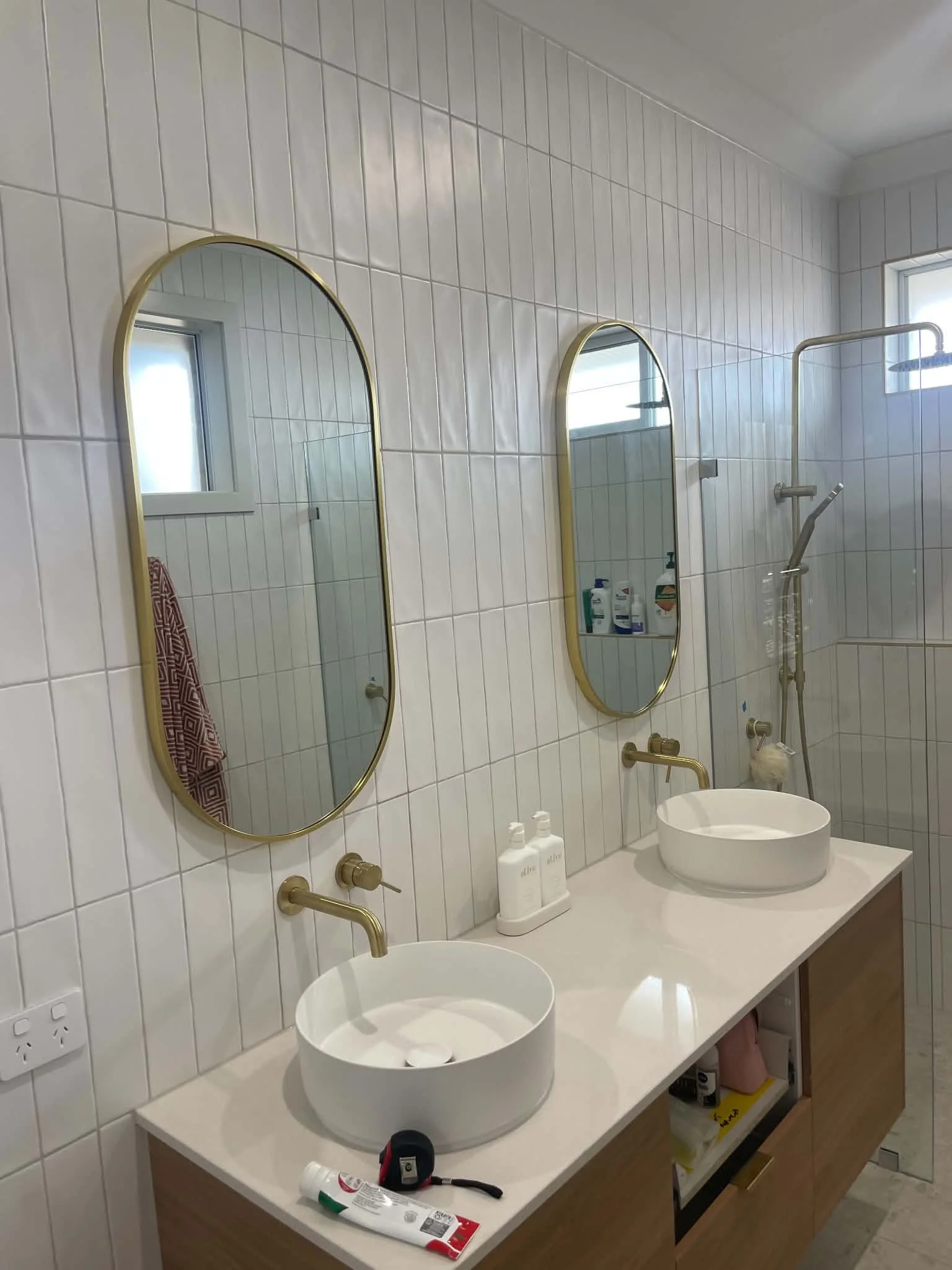 Modern bathroom with white tiled walls, two oval vessel sinks on a white countertop, and gold fixtures. There are two mirrors with gold frames above the sinks. A shower with a curved shower rod and a window in the shower area. Items on the countertop