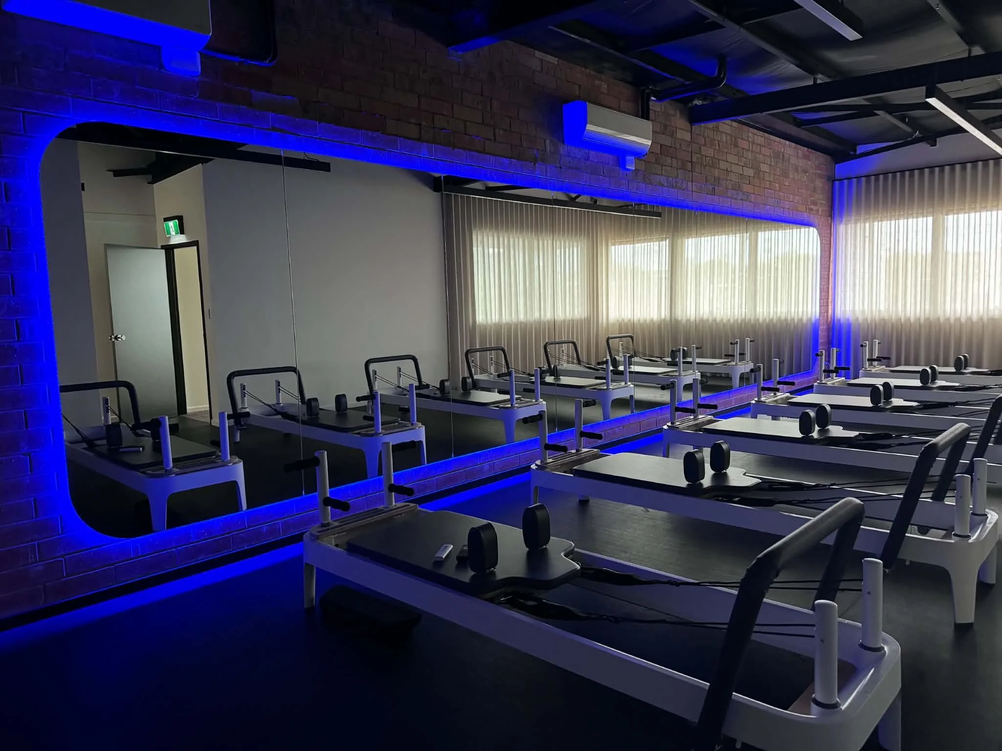 Fitness studio with Pilates reformer machines placed in rows, blue LED lighting along exposed brick walls, large window with curtains, and a mirror on one wall.