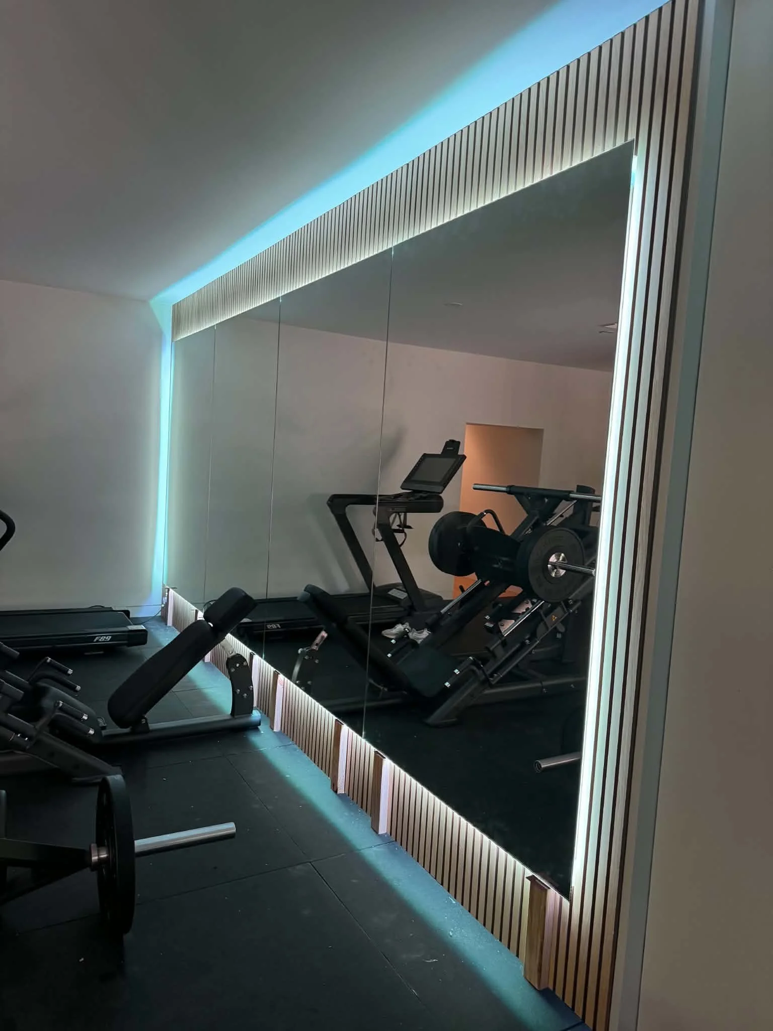A gym with dark flooring, a treadmill and exercise equipment facing a large mirror with a wooden slat border, illuminated with LED lighting.