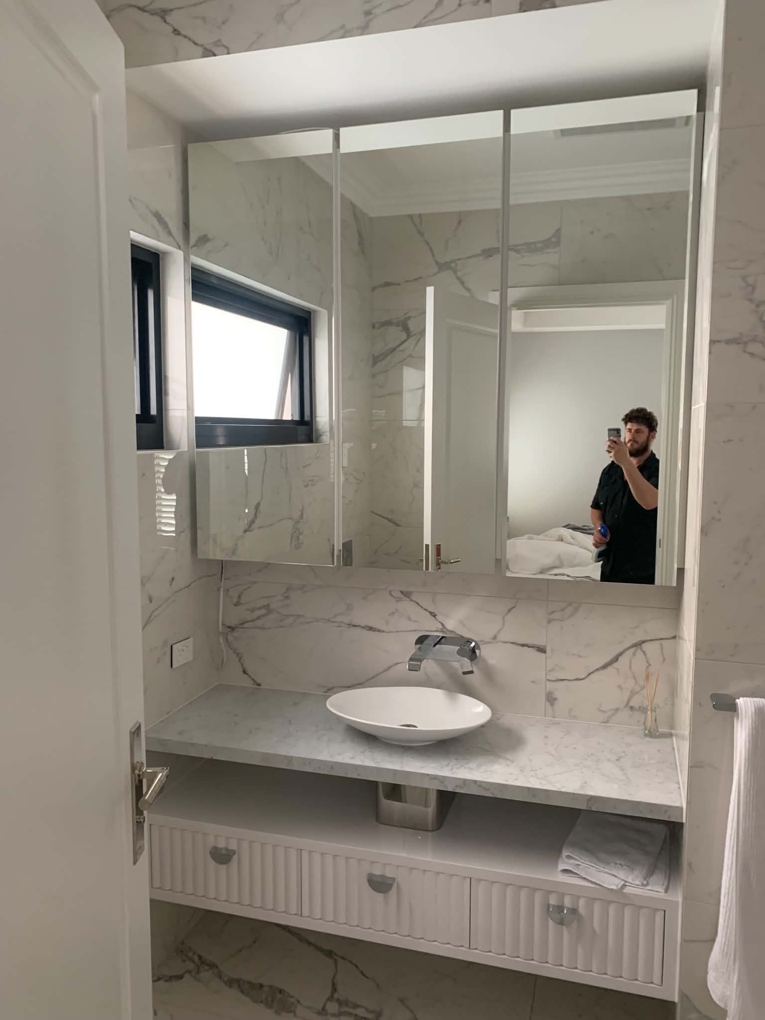 A bathroom vanity with drawers and a vessel sink, marble countertops and walls, and a large mirror reflecting a man taking a selfie. There is a towel and a washcloth visible.