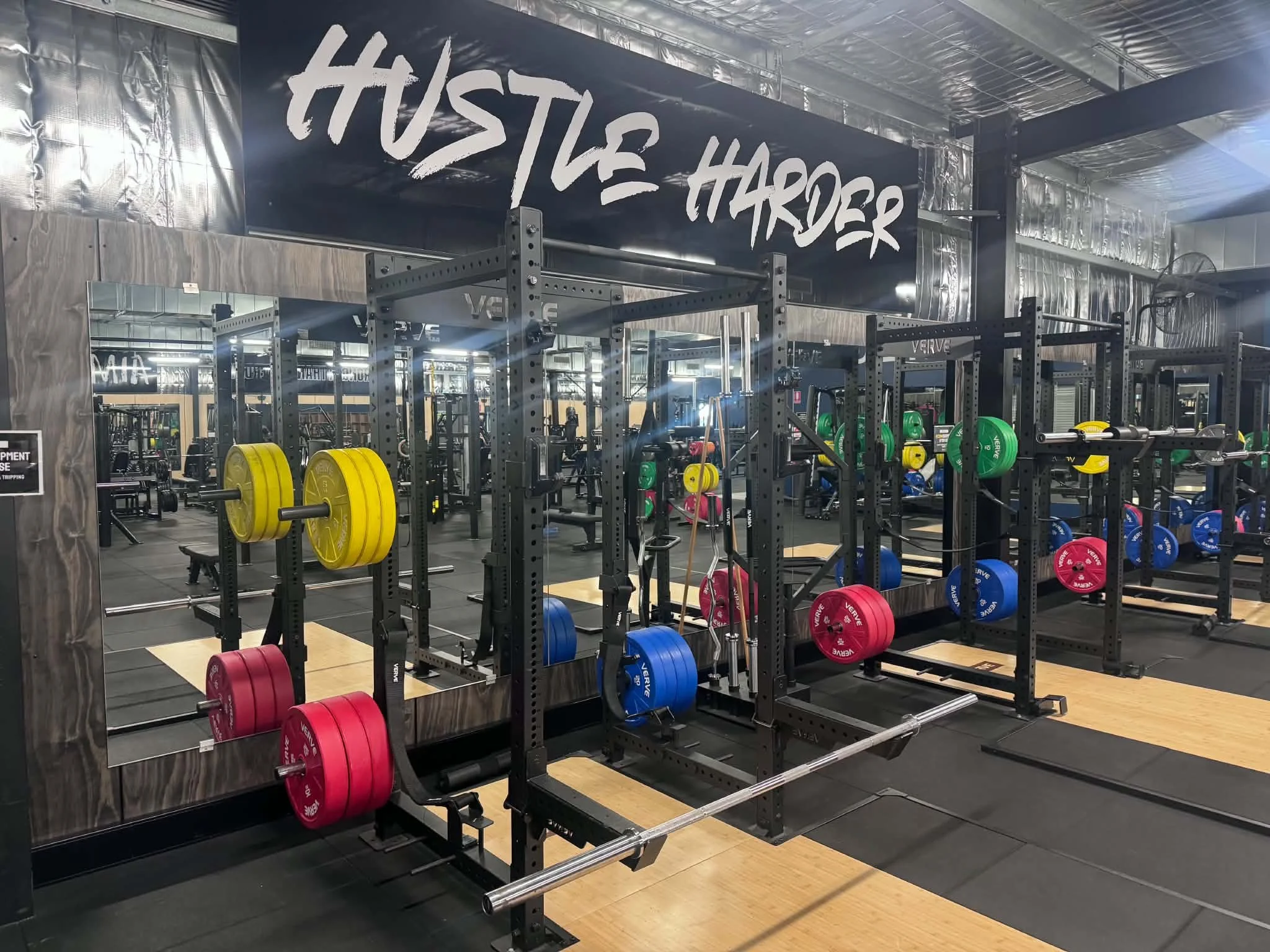 Gym weightlifting equipment with large mirrors on walls with colorful weights, black metal racks, and a large sign that reads 'HUSTLE HARDER'.