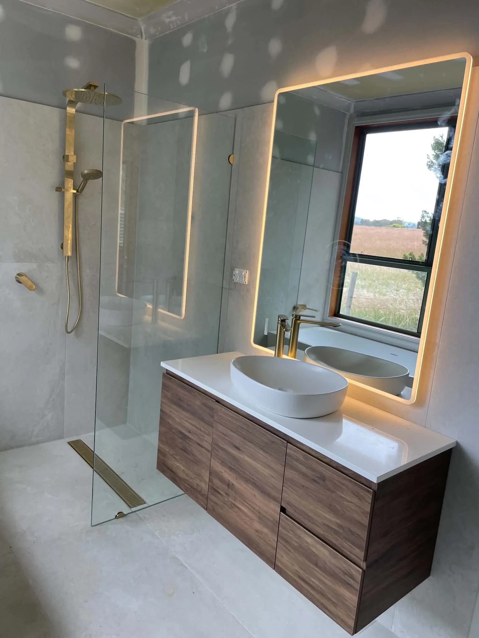 Modern bathroom with a large mirror with LED lighting, a white vessel sink on a wooden vanity, a window showing an outdoor view, and a shower area with a glass partition and a rain shower head.