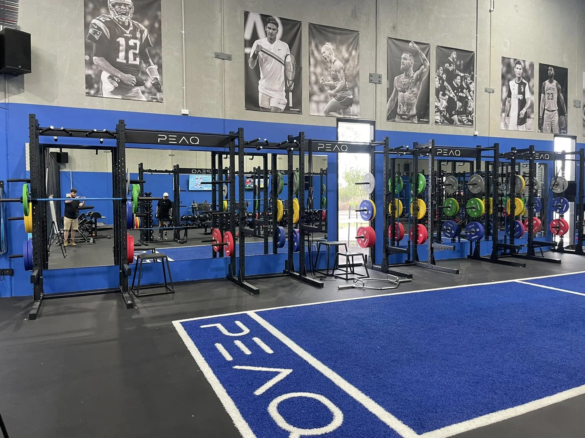 View of a gym with black weightlifting racks, colorful weight plates, and people lifting weights. Large black and white posters of famous athletes on the wall.