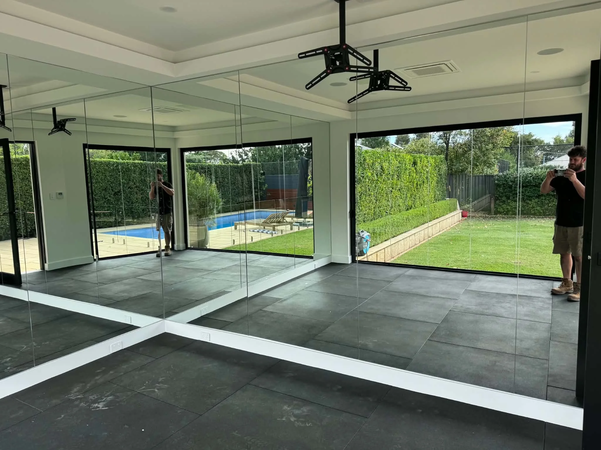 A modern interior room with large mirrored walls, dark tiled floor, and ceiling-mounted black clothing racks. Outside the large windows, a backyard with a pool, green lawn, and trees is visible.