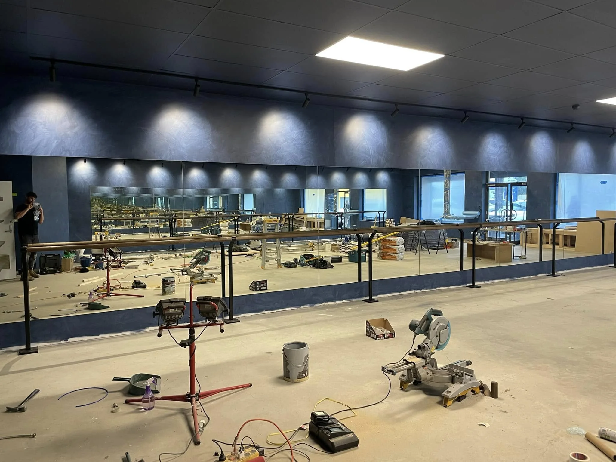 Interior of a building under construction with tools, equipment, and materials scattered on the floor, partially finished walls with a large mirror, and a ceiling with bright lighting.