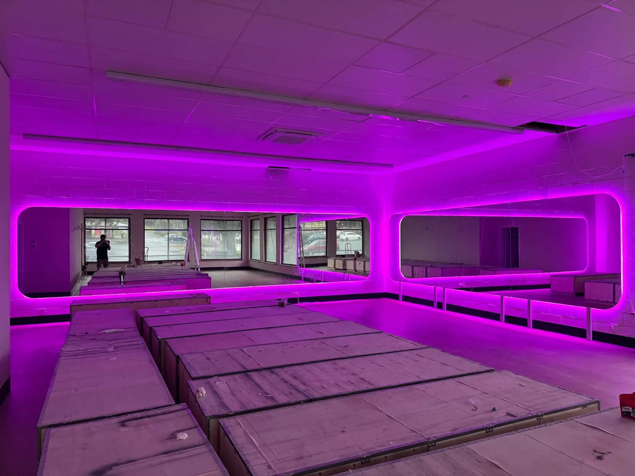 An empty room with purple LED lighting, large mirrors on the wall, ceiling tiles, and multiple wooden pallets stacked in rows. A person is visible in the reflection of the mirrors, standing near the windows.
