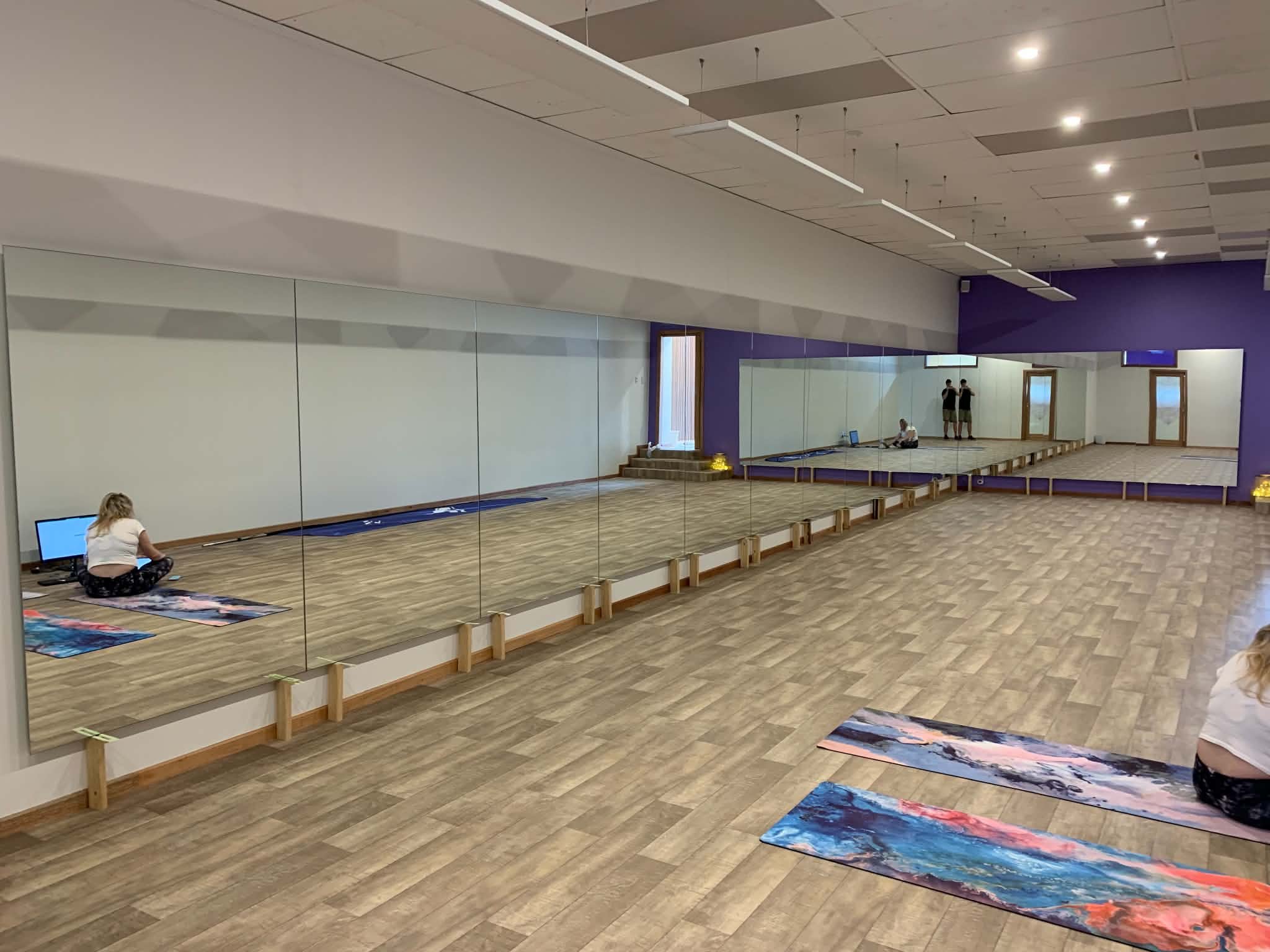 A large, empty dance studio with wooden floors, a mirrored wall, and purple walls. There are yoga mats on the floor, and a few people are sitting or standing in the room. The ceiling has white panels with several lights, and there are windows at the 