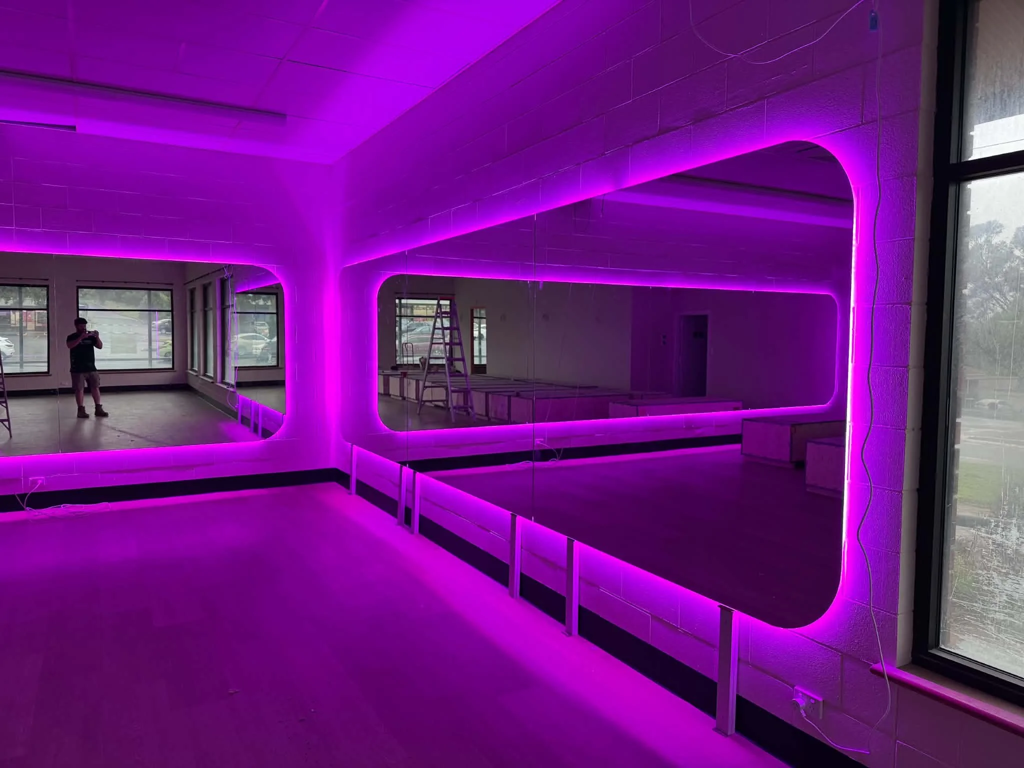 A room with neon purple lighting along the ceiling and walls behind a large mirror, large mirrors, windows, a ladder.