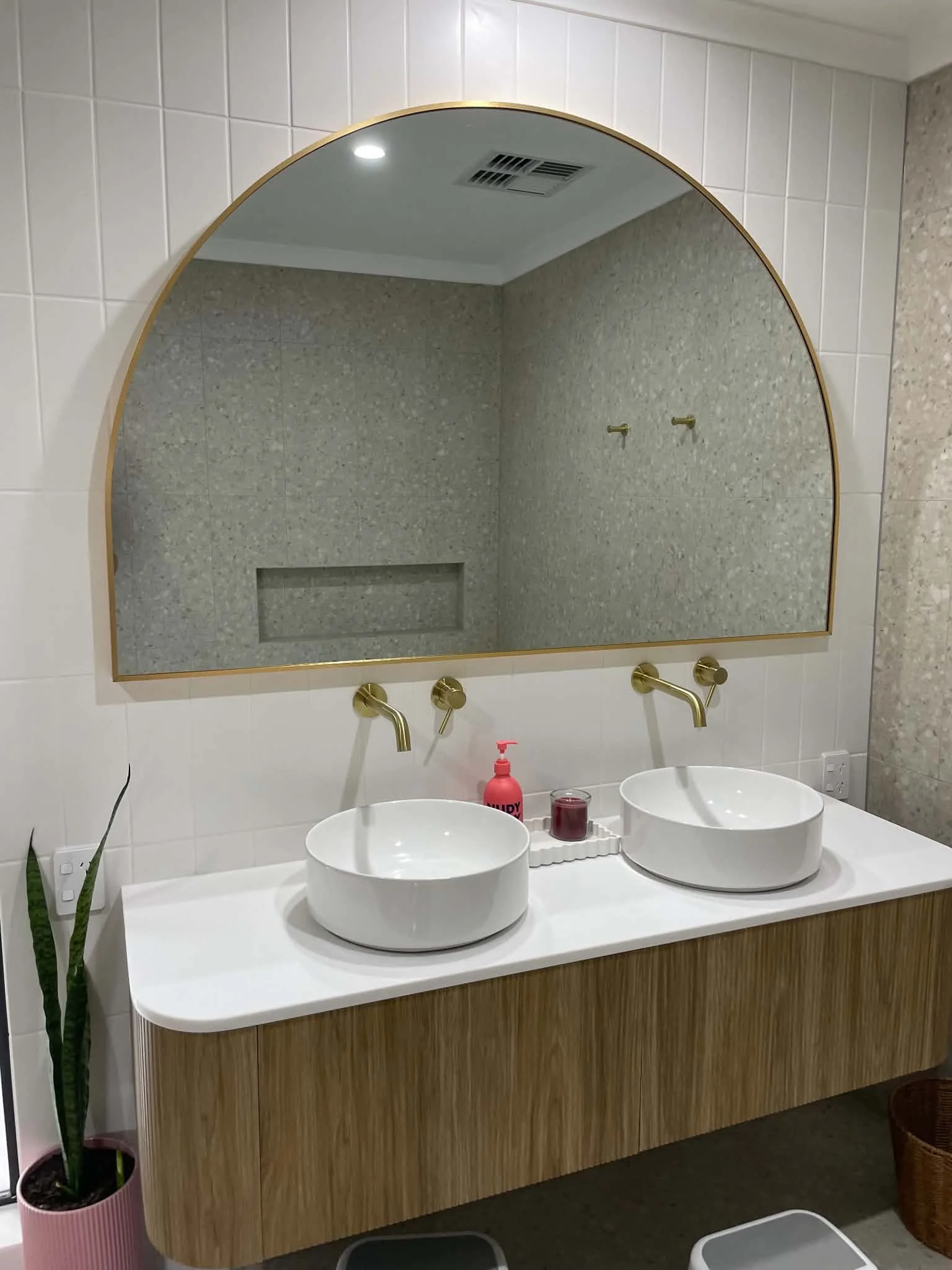 Bathroom vanity with two white vessel sinks, gold faucets, a large arched mirror with a gold frame, and a countertop with soap and a small candle. A potted plant is visible on the left, and there is a wicker basket on the floor to the right.