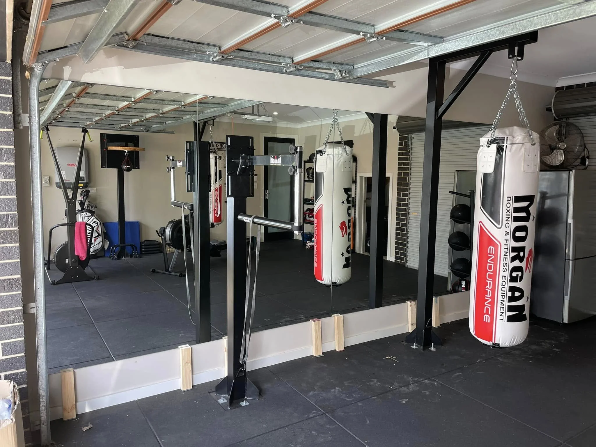 A home gym setup with boxing and fitness equipment, including a punching bag, a speed bag, suspension trainer, and a mirrored wall in a garage space.