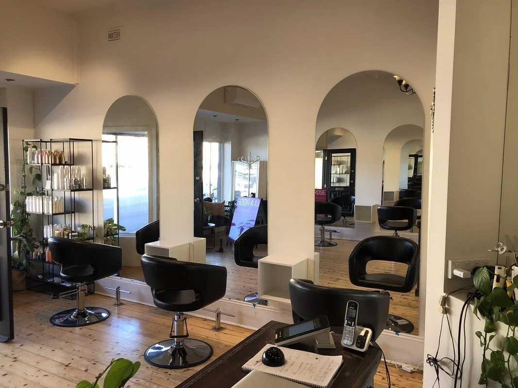 Interior of a hair salon with three black styling chairs in front of large mirrors, a shelving unit with hair products and plants, wooden flooring, and natural light coming through windows.