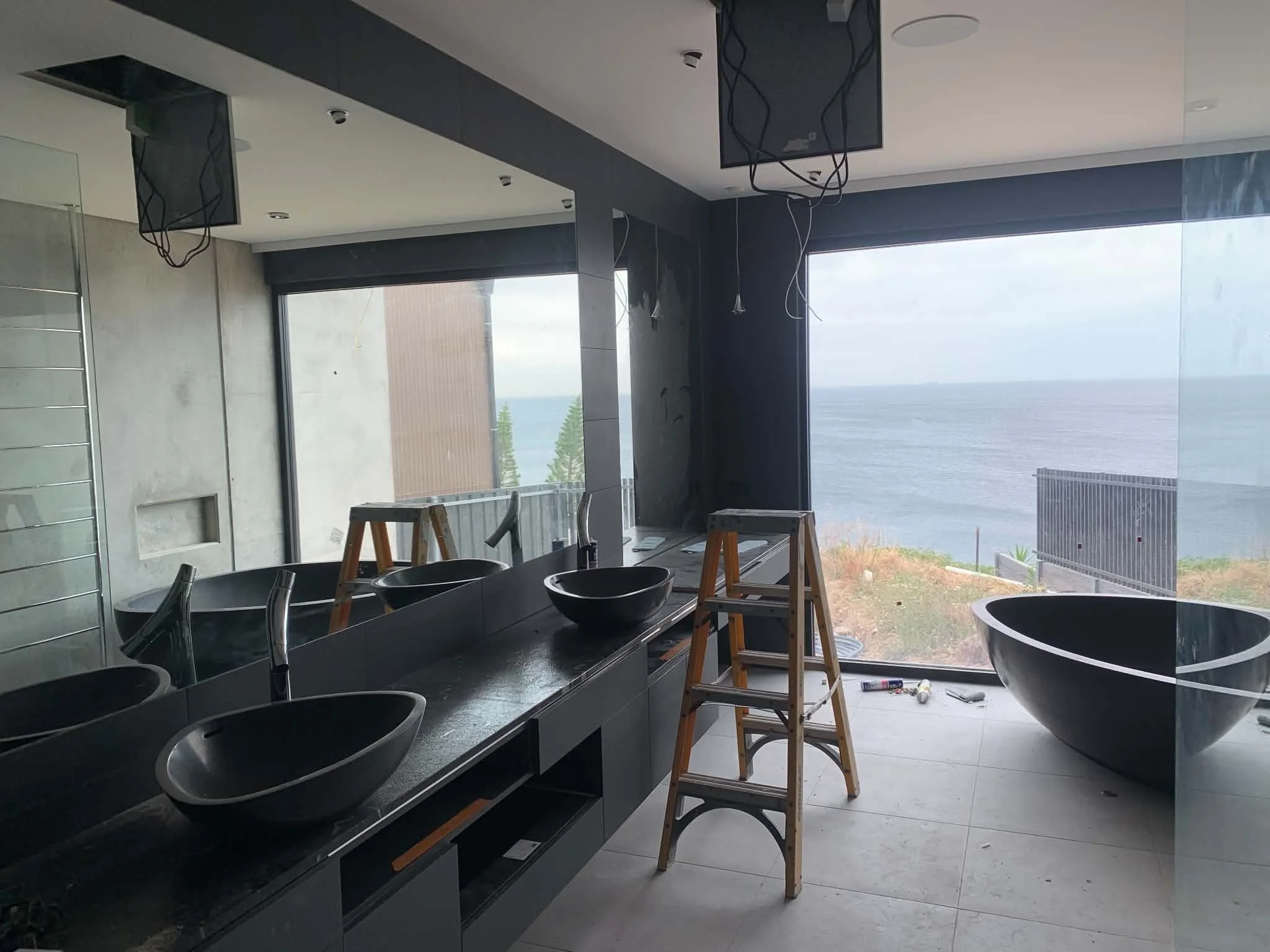 The image shows a modern bathroom under construction with black sinks, a large window overlooking an ocean, and construction tools and ladders present, indicating ongoing installation or renovation work.