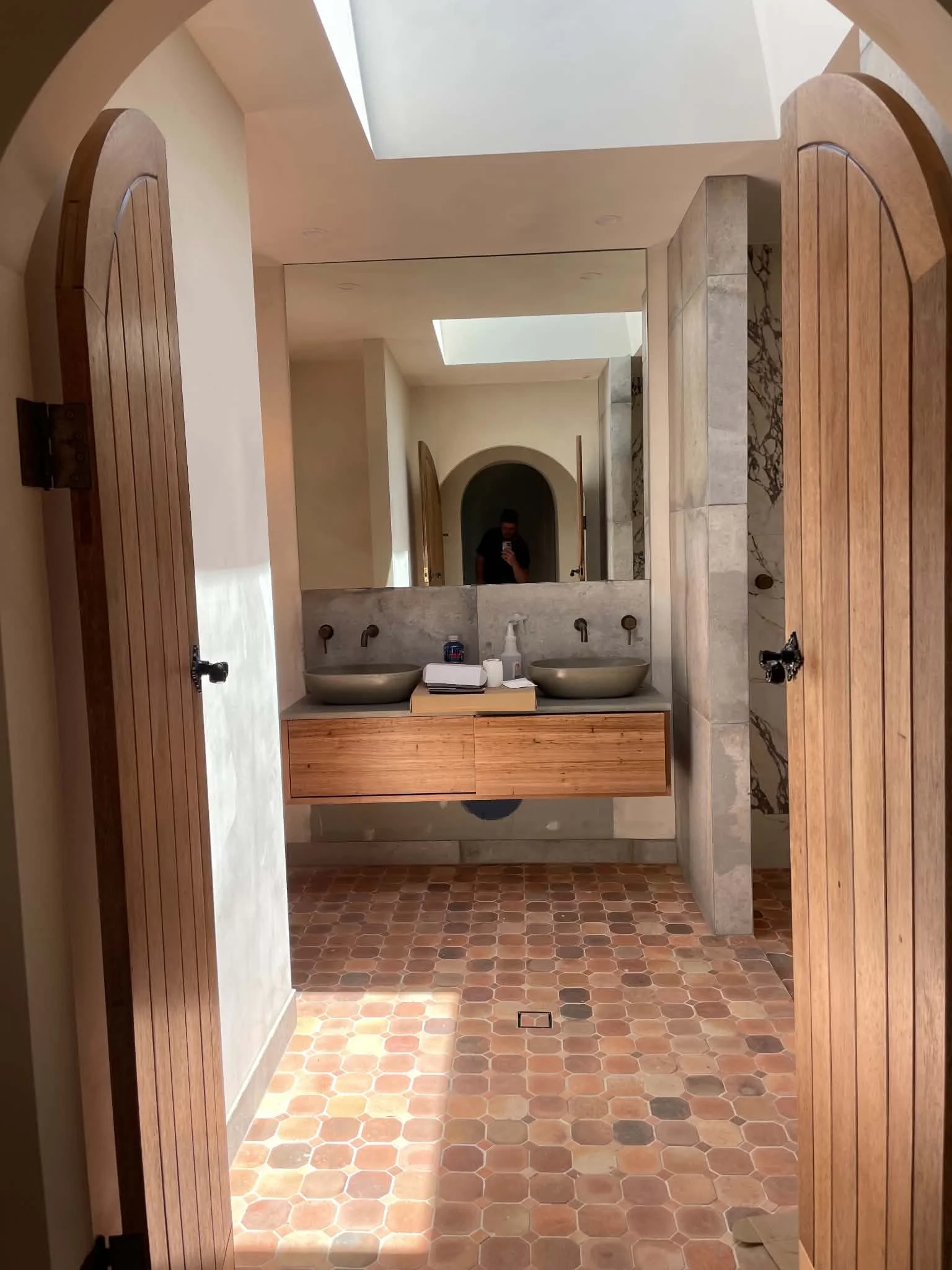 A bathroom with a stone and wood vanity featuring two vessel sinks and a large mirror. The floor is covered with small hexagonal terracotta tiles, and there are skylights in the ceiling providing natural light.