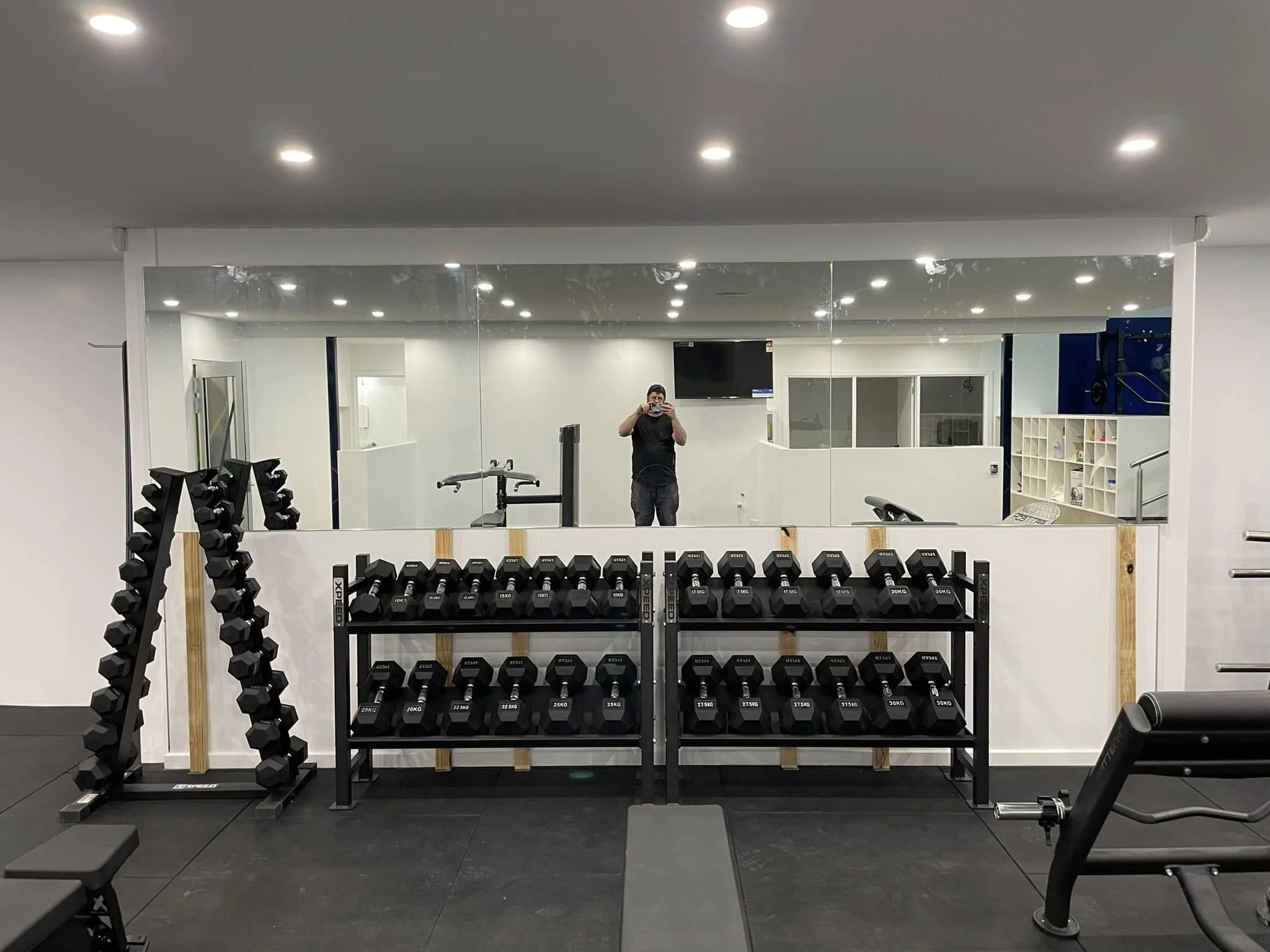 A mirror showing a man taking a photo in a home gym, with dumbbells on racks in the foreground, exercise benches, a TV, and shelving in the background.