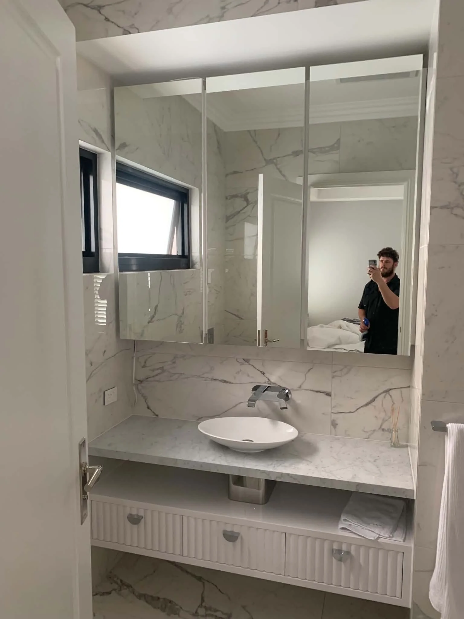 A bathroom with marble walls and a large mirror. The mirror reflects a person taking a selfie, a window, and part of the room beyond. The bathroom has a modern sink on a marble countertop, and a white cabinet underneath with small drawers. There is a towel folded on the cabinet and a tissue box on the right side.