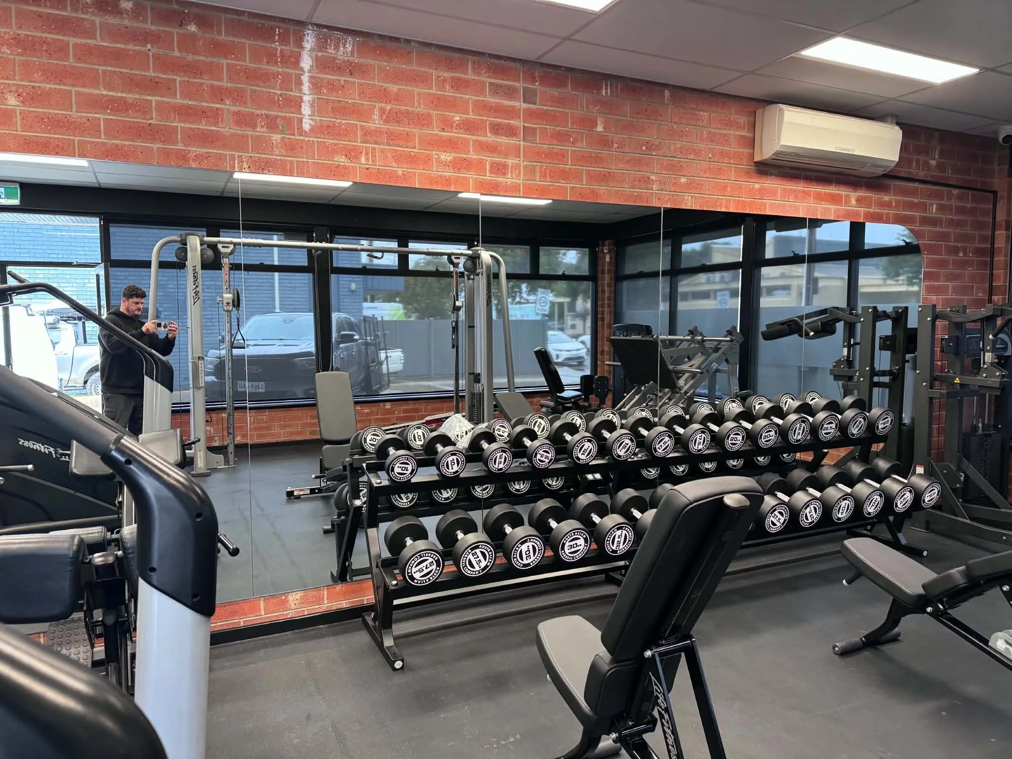 A gym with workout equipment including dumbbells on racks, a treadmill, and various exercise machines. Large windows show parked cars outside. 
