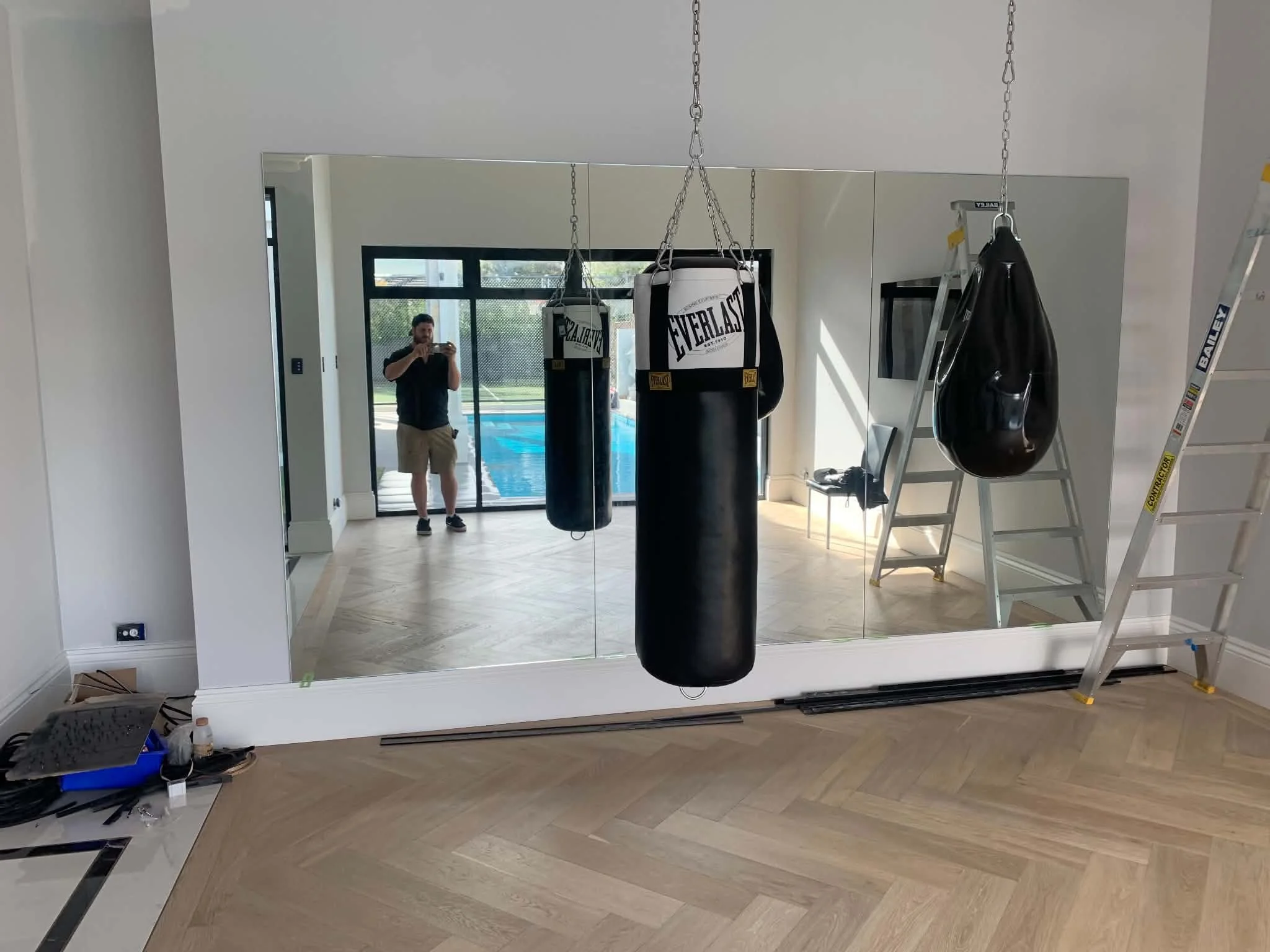 The image shows a room with large mirror sliding doors. In front of the mirror, there are boxing punching bags hanging from the ceiling. A ladder and some boxing gloves are also visible. A person is taking a photo of the room from the reflection of t