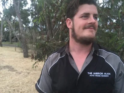 A man with dark hair and a beard, wearing a black and gray shirt, standing outdoors in top that says  Mirror Man Adelaide in print