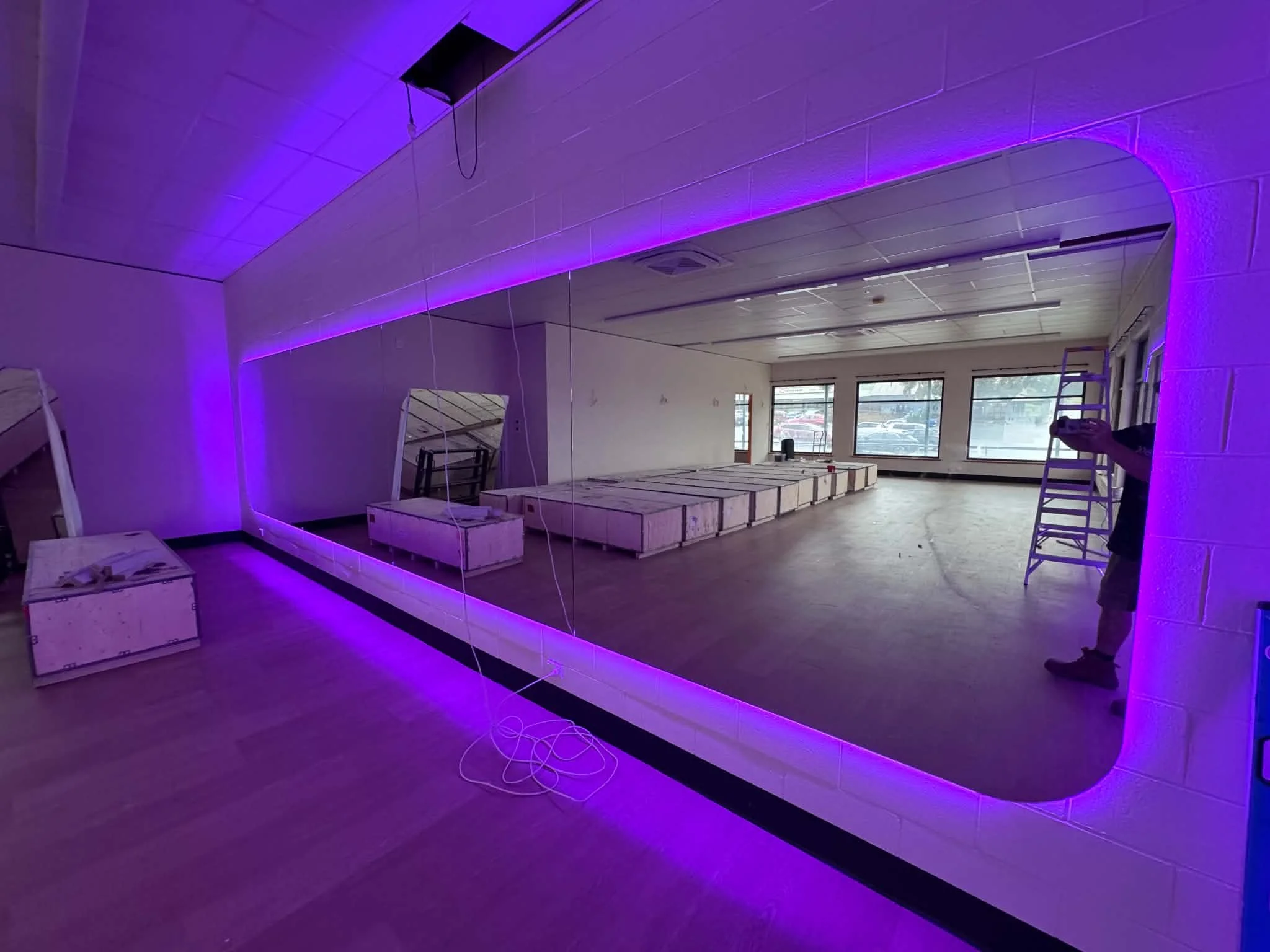 An interior room under construction with purple LED strip lighting along a curved ceiling. There are wooden crates, ladders, and a person on the right side, partially visible.