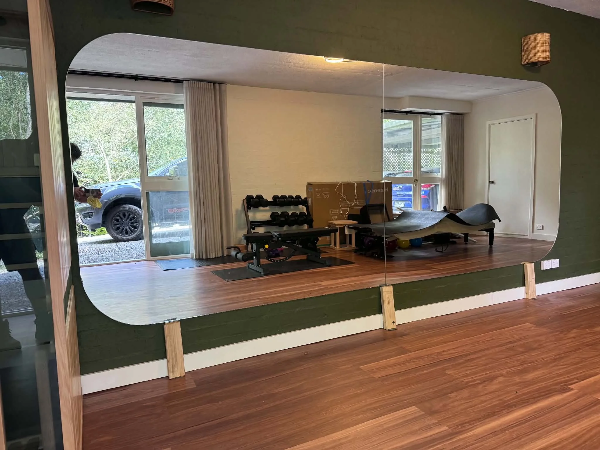Large mirror reflecting a room with two windows, gym equipment, and a person taking a photo with a phone.