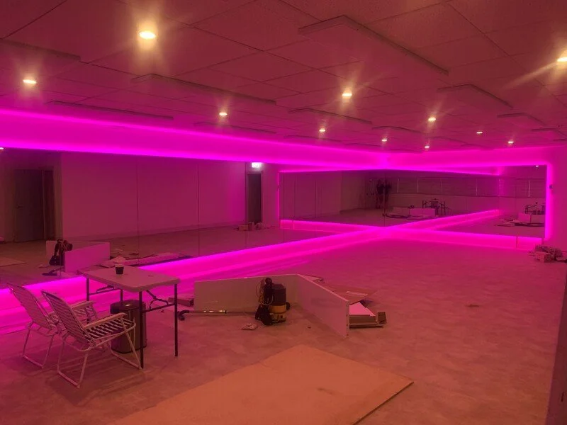 Interior of a room under construction with pink neon lighting along the walls, mirrors covering one wall, and construction tools and furniture scattered on the floor.