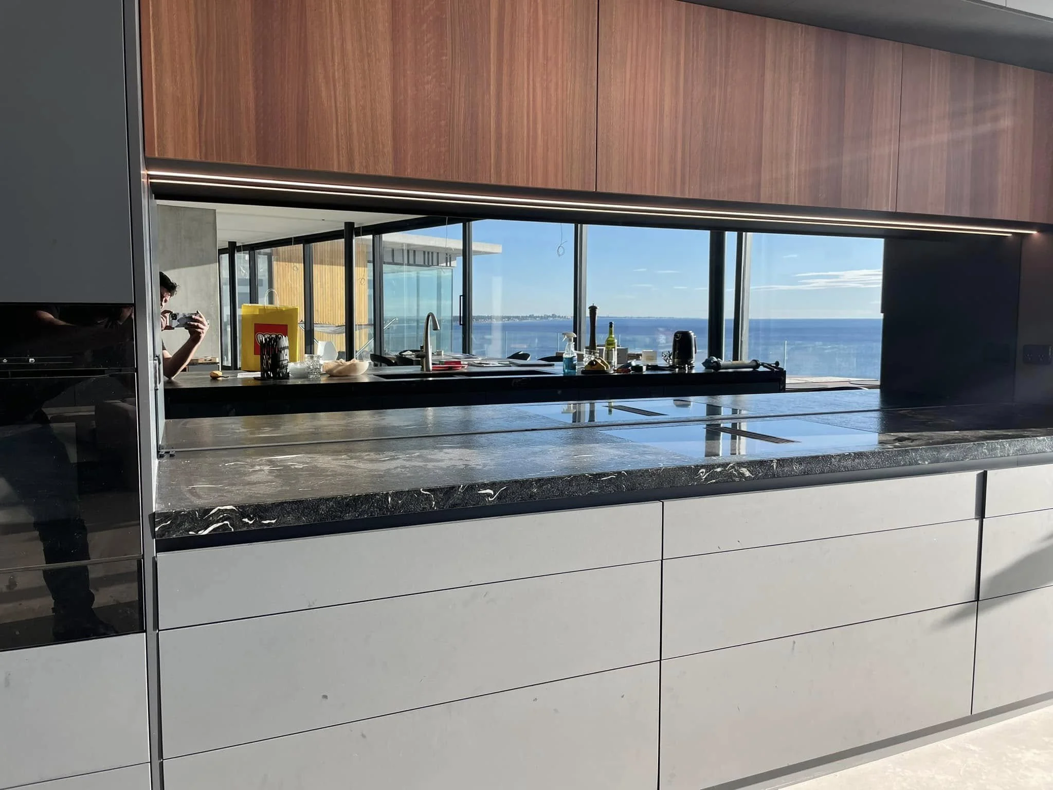 Modern kitchen with large windows overlooking a body of water, wooden wall cabinets, and a black countertops with various kitchen items.