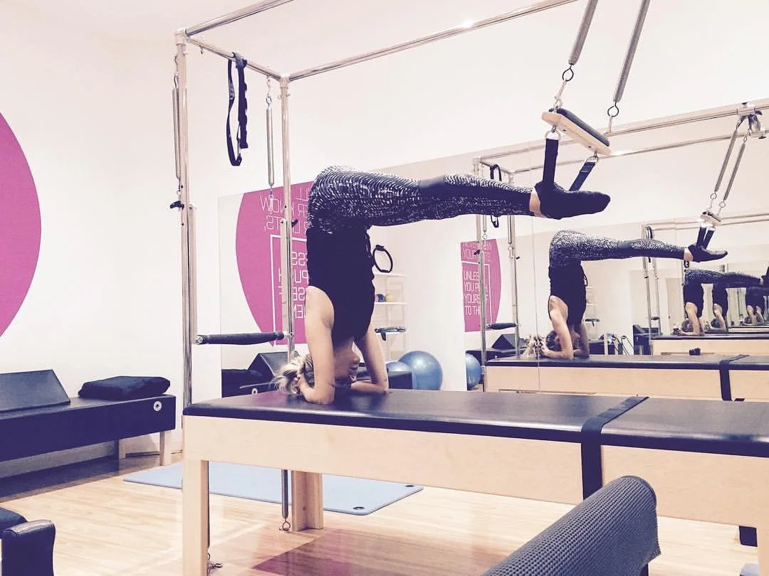 A person practicing yoga on a reformer machine in a fitness studio, with mirrors aligned to help technique, performing a headstand pose with legs extended backward.