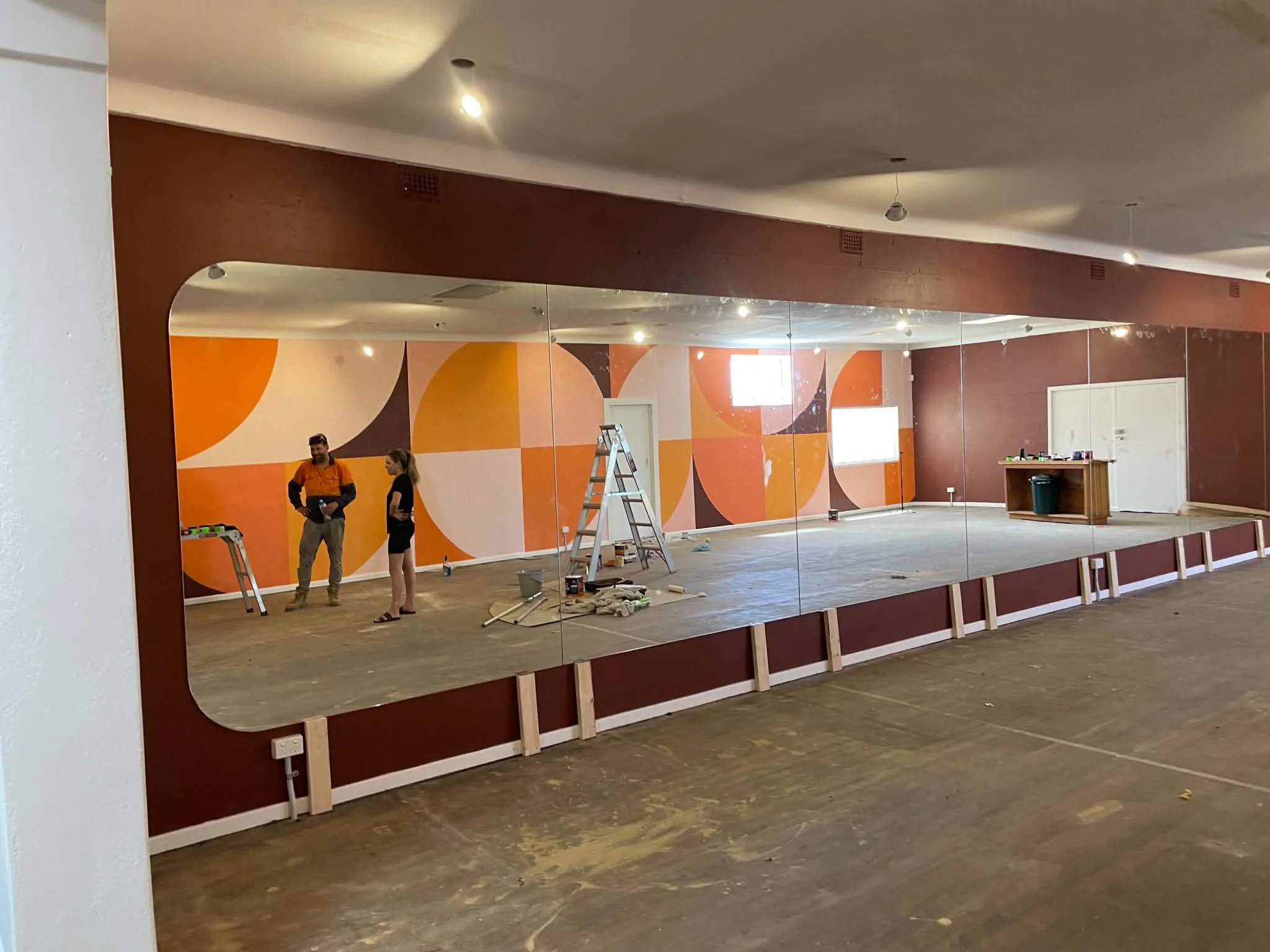 Large mirror reflecting an indoor room with orange and beige abstract wall art, people standing near a ladder, and various tools on the floor, suggesting an ongoing renovation or decorating project.