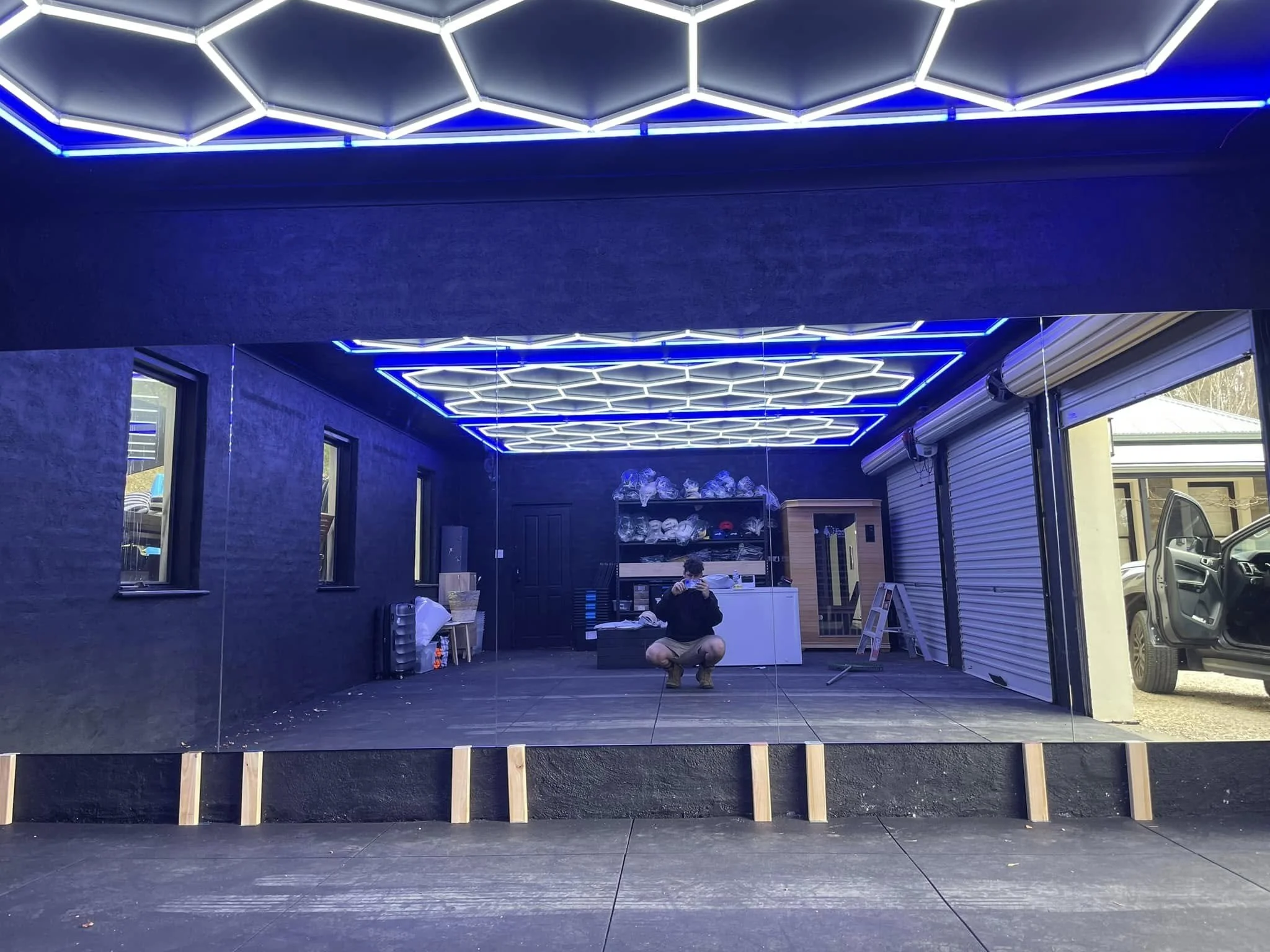Reflective glass window showing a person squatting and taking a photo inside a room with blue neon hexagonal ceiling lights, dark walls, and open garage door.