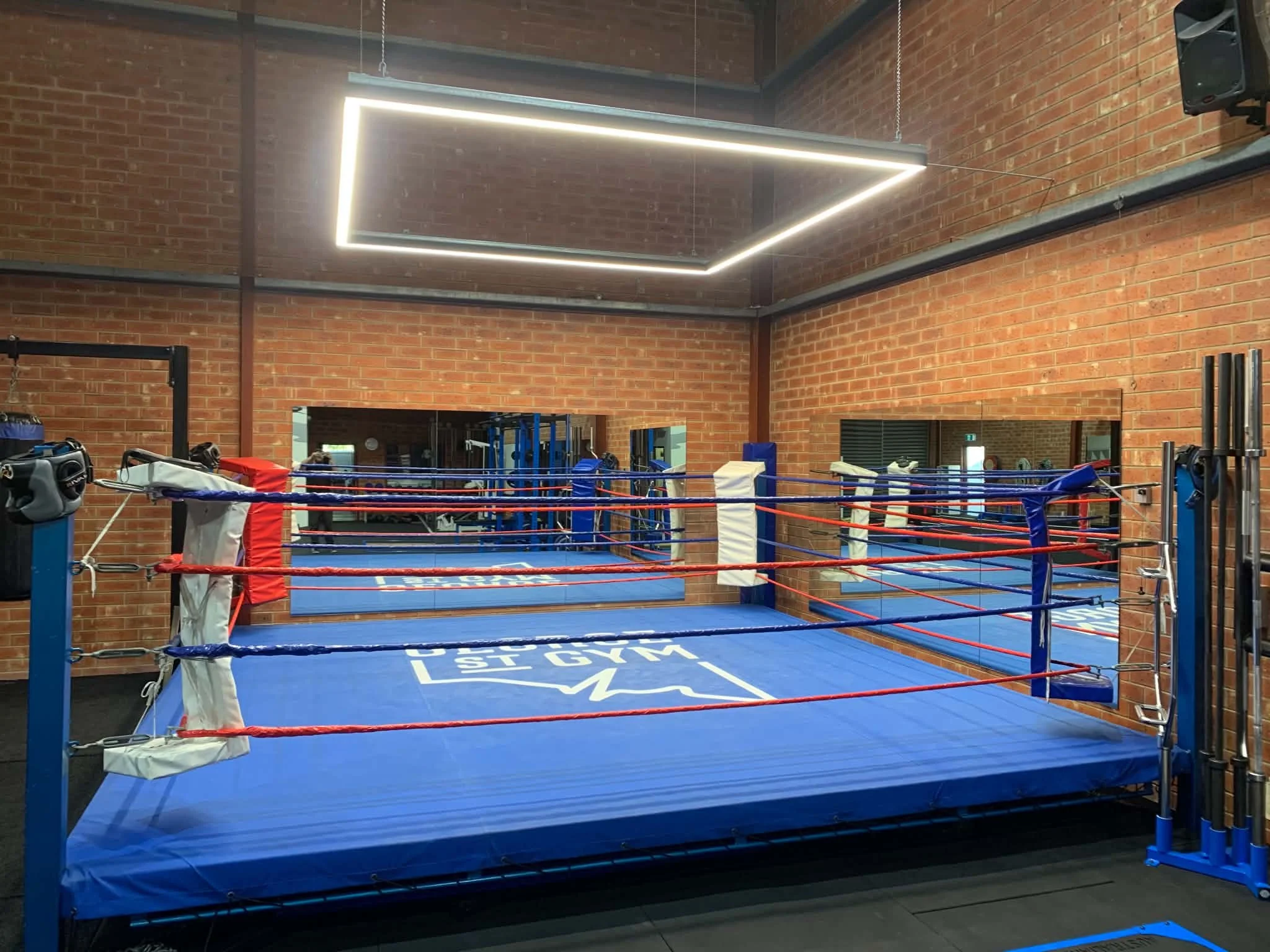 Indoor boxing gym with a boxing ring featuring blue padding, red and blue ropes, and a large illuminated rectangular mirror on the brick wall