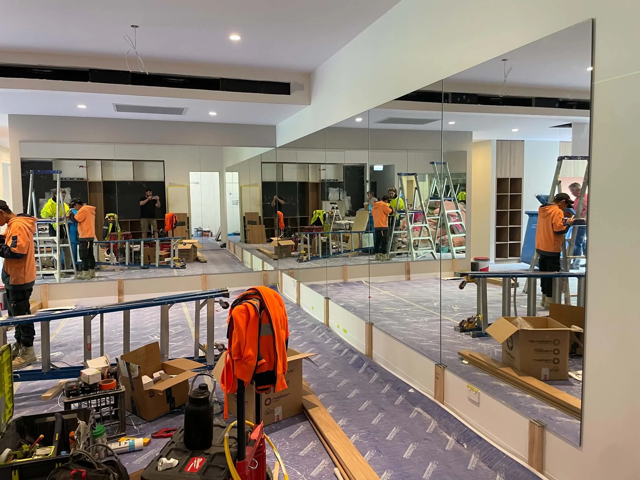 Interior of a building under construction or renovation, with workers installing mirrors on the wall, ladders, and various tools and boxes scattered around.
