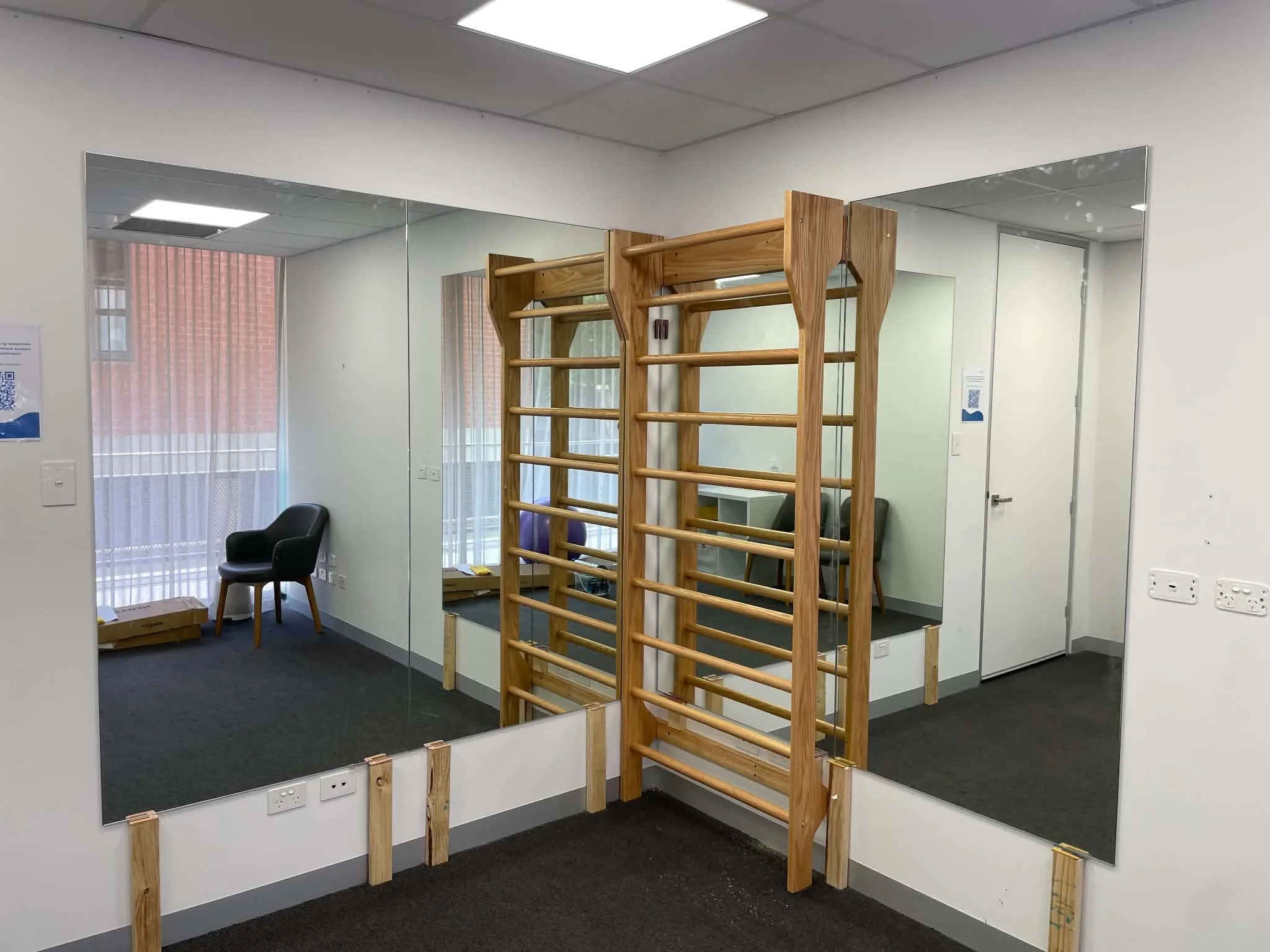 An office room with mirror walls, a wooden climbing frame, and a black chair in the corner.