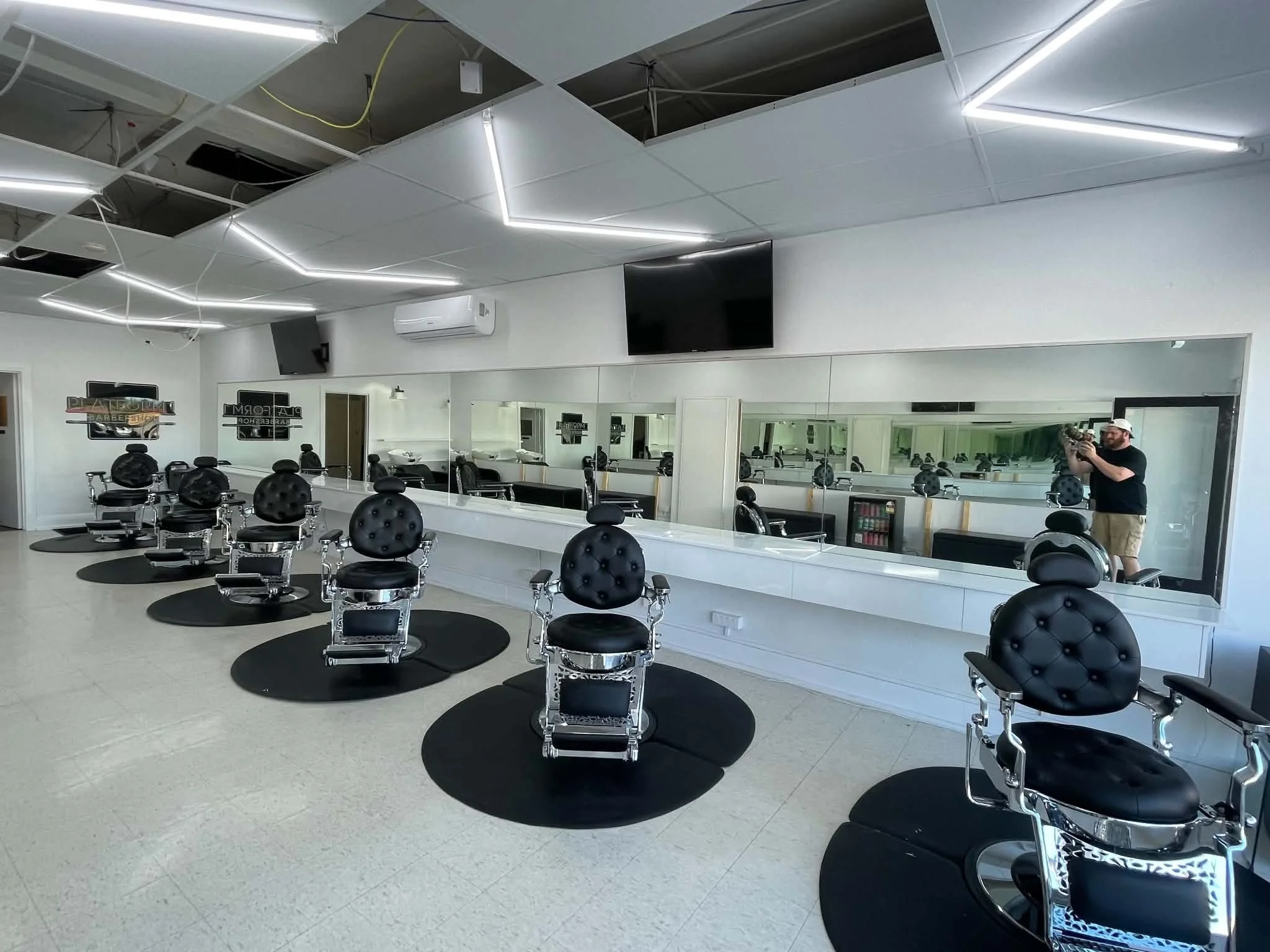 Interior of a hair salon with multiple black barber chairs arranged in front of large mirrors, a wall-mounted TV, and modern lighting fixtures on the ceiling.