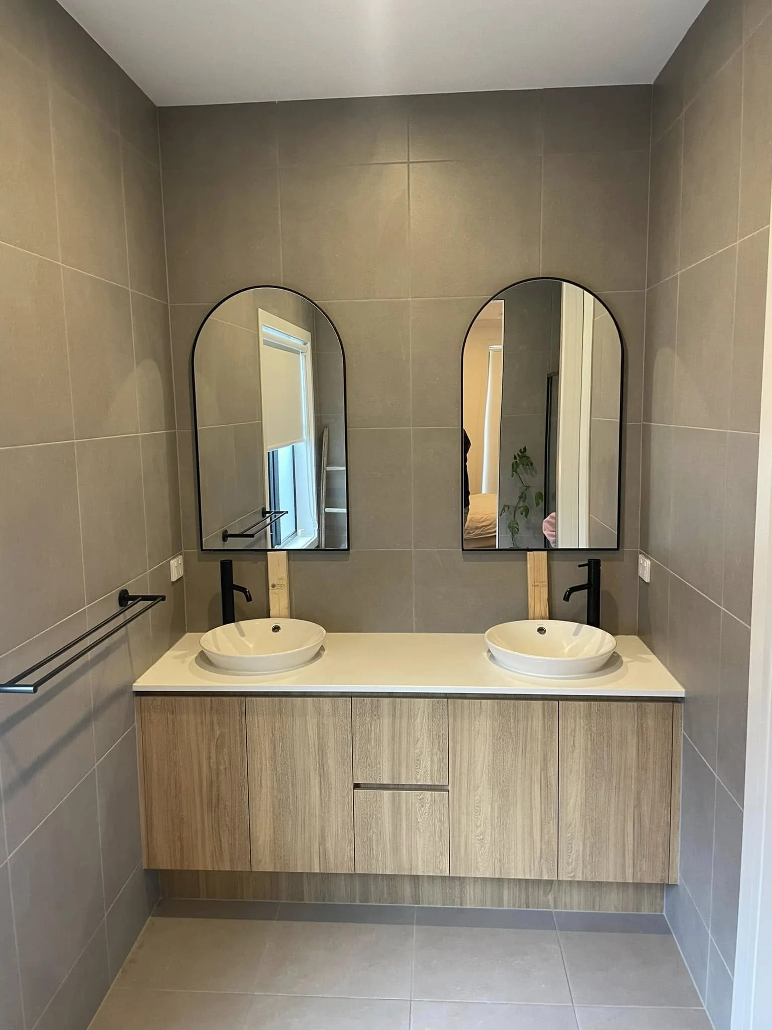 A modern bathroom with two vessel sinks on a wooden vanity, two arched mirrors above each sink, black faucets and towel bars, beige tiled walls, and a beige tiled floor.