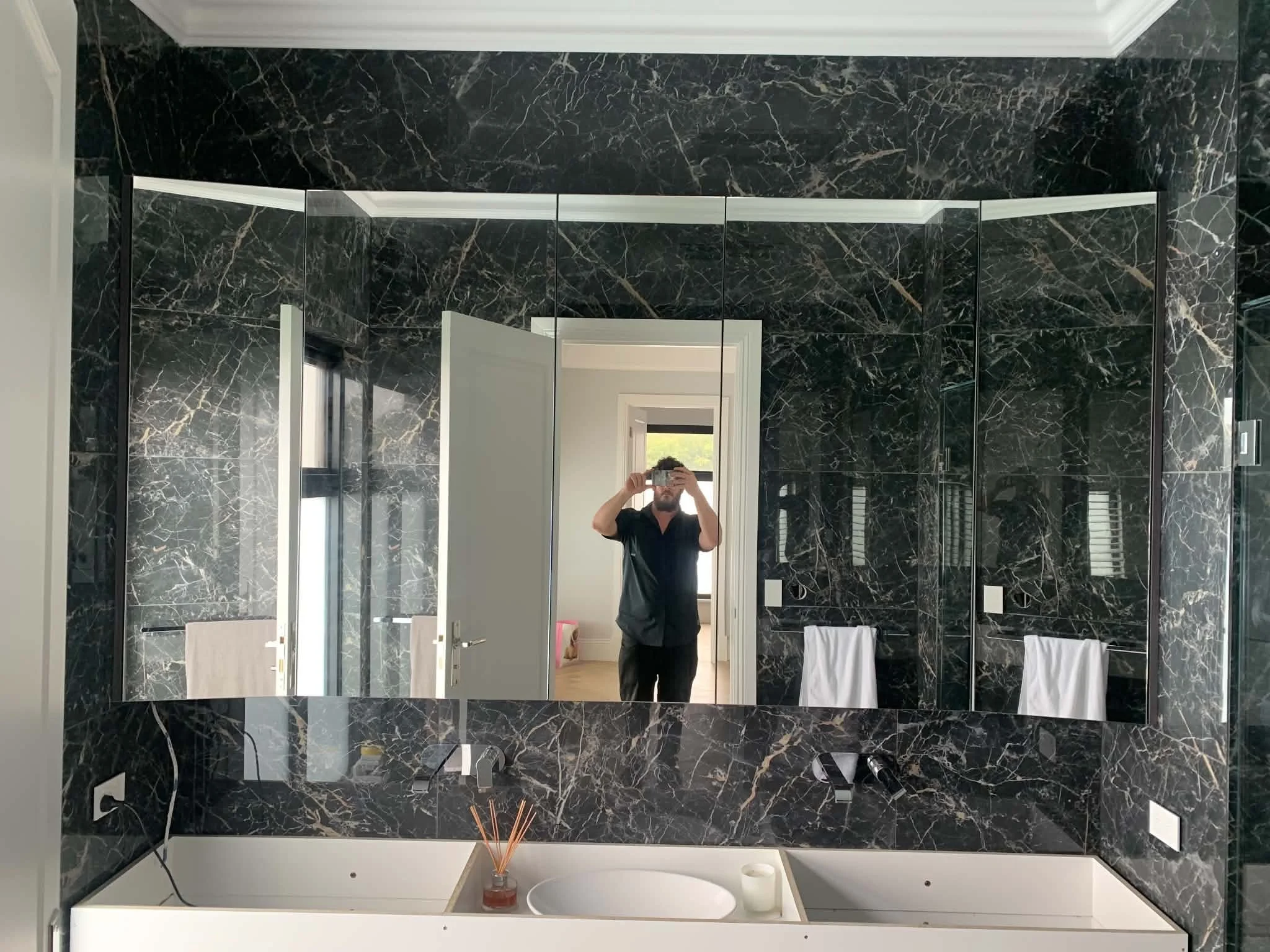 A bathroom with walls and a large mirror covered in black marble with white veining. The reflection shows a doorway to another room, a person taking the photo with a smartphone, and some bathroom fixtures. The ceiling has decorative white crown moldi