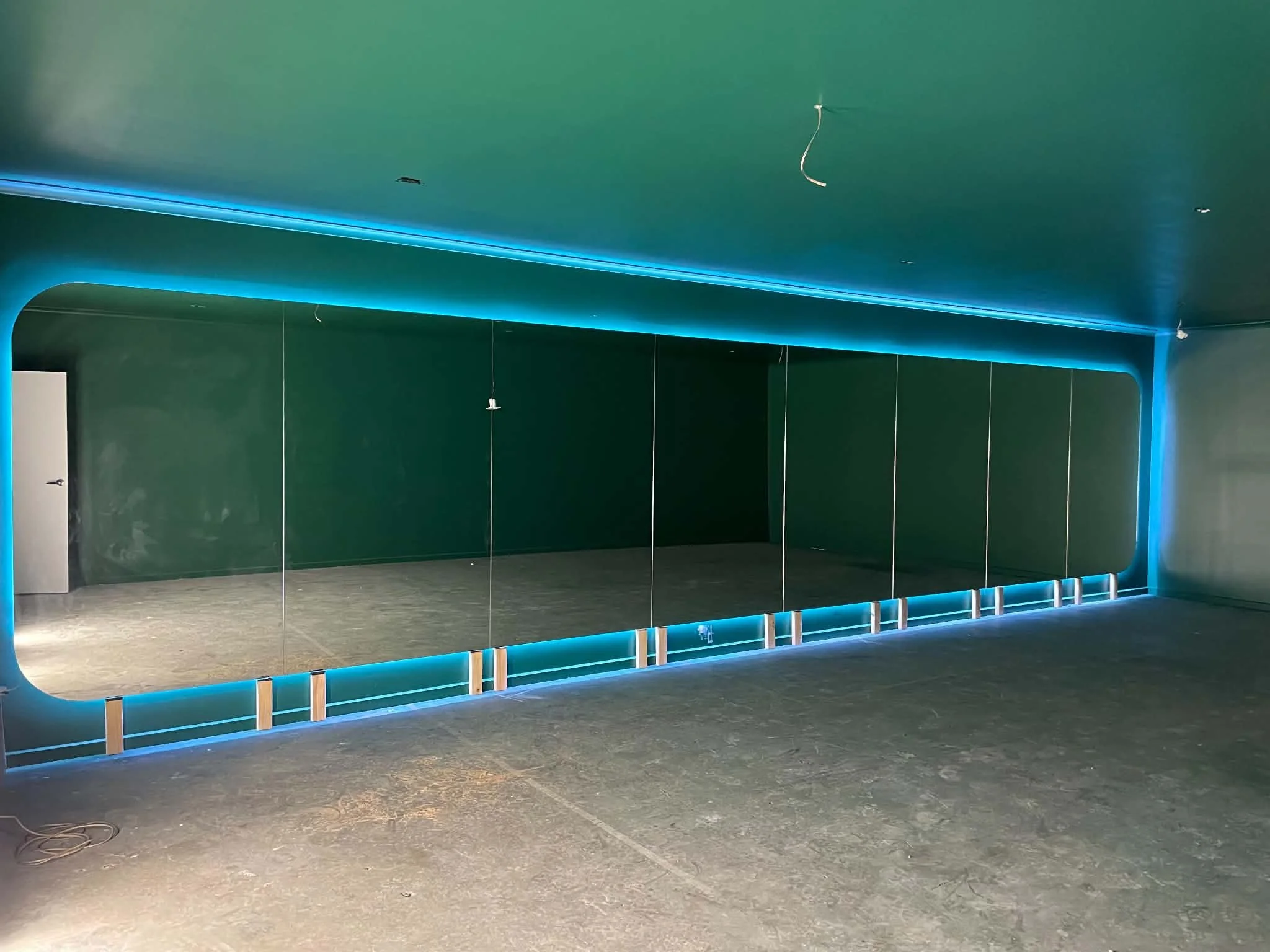 An indoor space with a dark green wall and ceiling, illuminated by blue LED strip lighting along the ceiling and floor. There is a partition with frosted glass panels, and some electrical wires hanging from the ceiling, possibly for lighting or fixtu