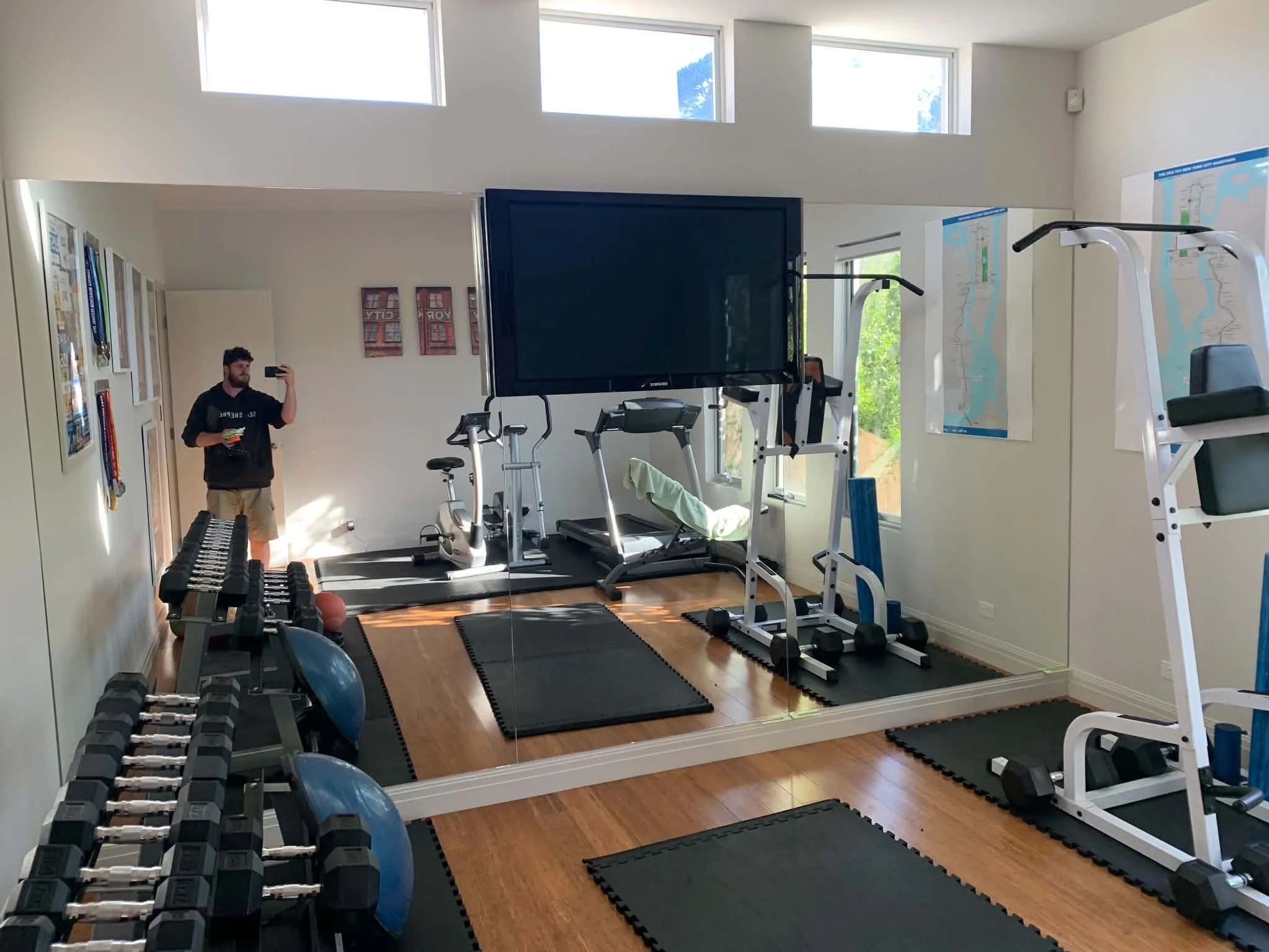 A home gym with various workout equipment including dumbbells, a treadmill, stationary bikes, a weight machine, and a large television. Sunlight streams in through multiple windows, illuminating the room.