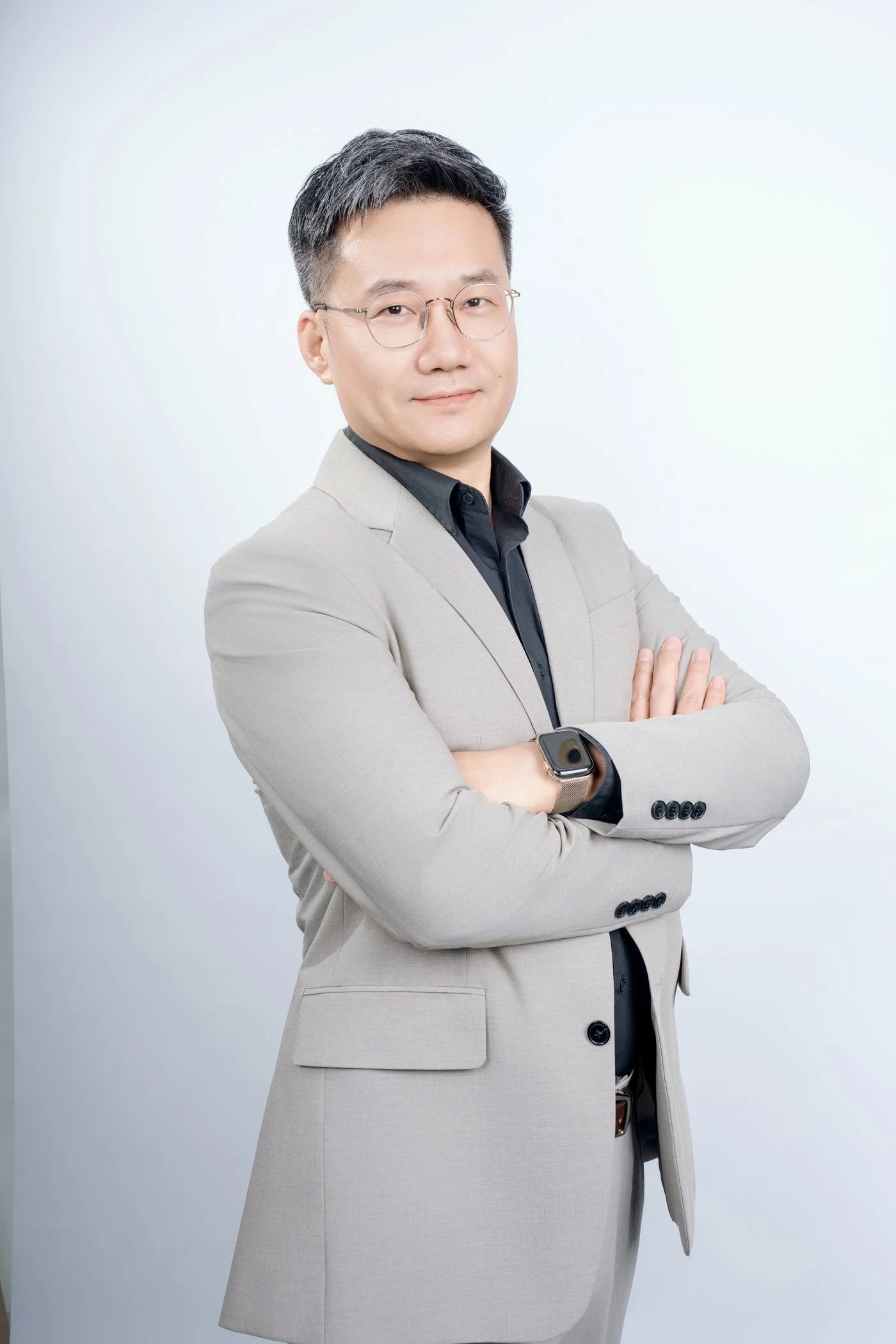회색 정장 차림의 남성이 팔짱을 끼고 미소를 짓고 있으며, 배경은 흰색이다.