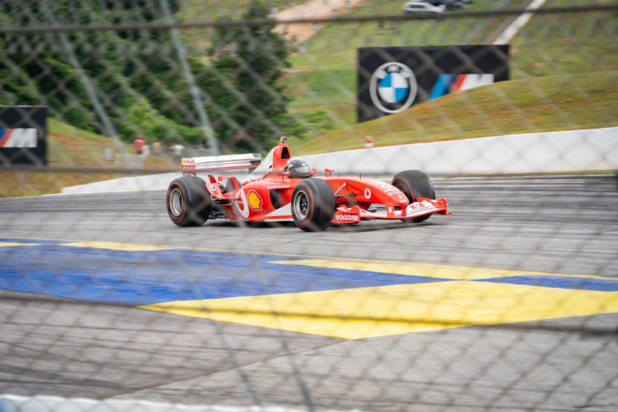 A red Formula 1 race car speeding on a race track with a fence in the foreground and blurred background of trees and drifted leaves.