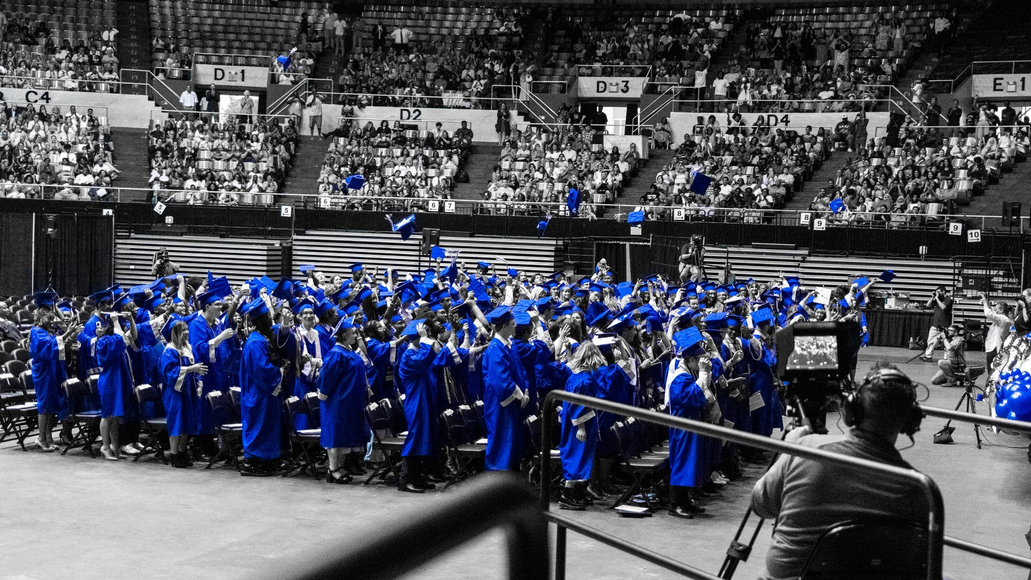 A graduation ceremony with students in blue caps and gowns celebrating in a large indoor arena filled with seated audience.