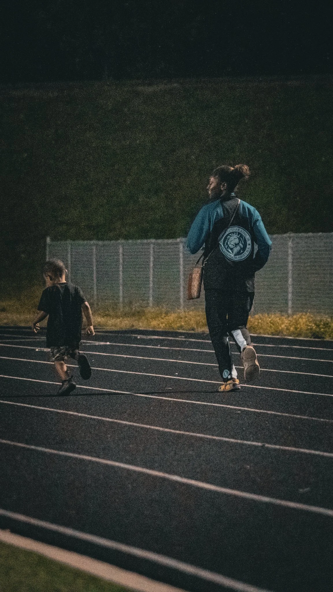 A woman and a young boy running on a track at night. The woman is wearing athletic clothing and a backpack, while the boy is dressed in shorts and a t-shirt.