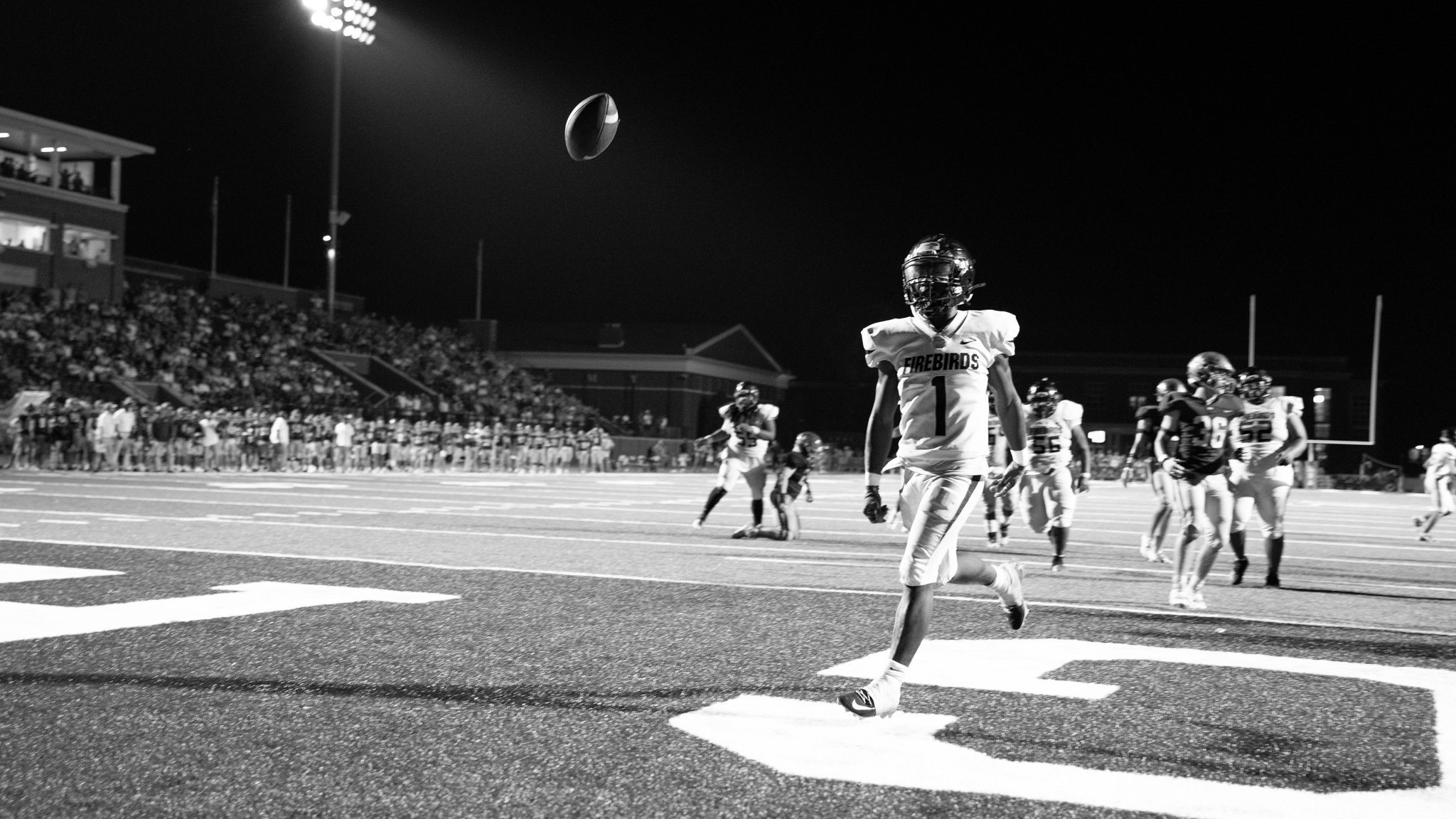 Nighttime football game with players on the field and crowd in the stands, player in jersey number 1 walking near sideline, football in midair.