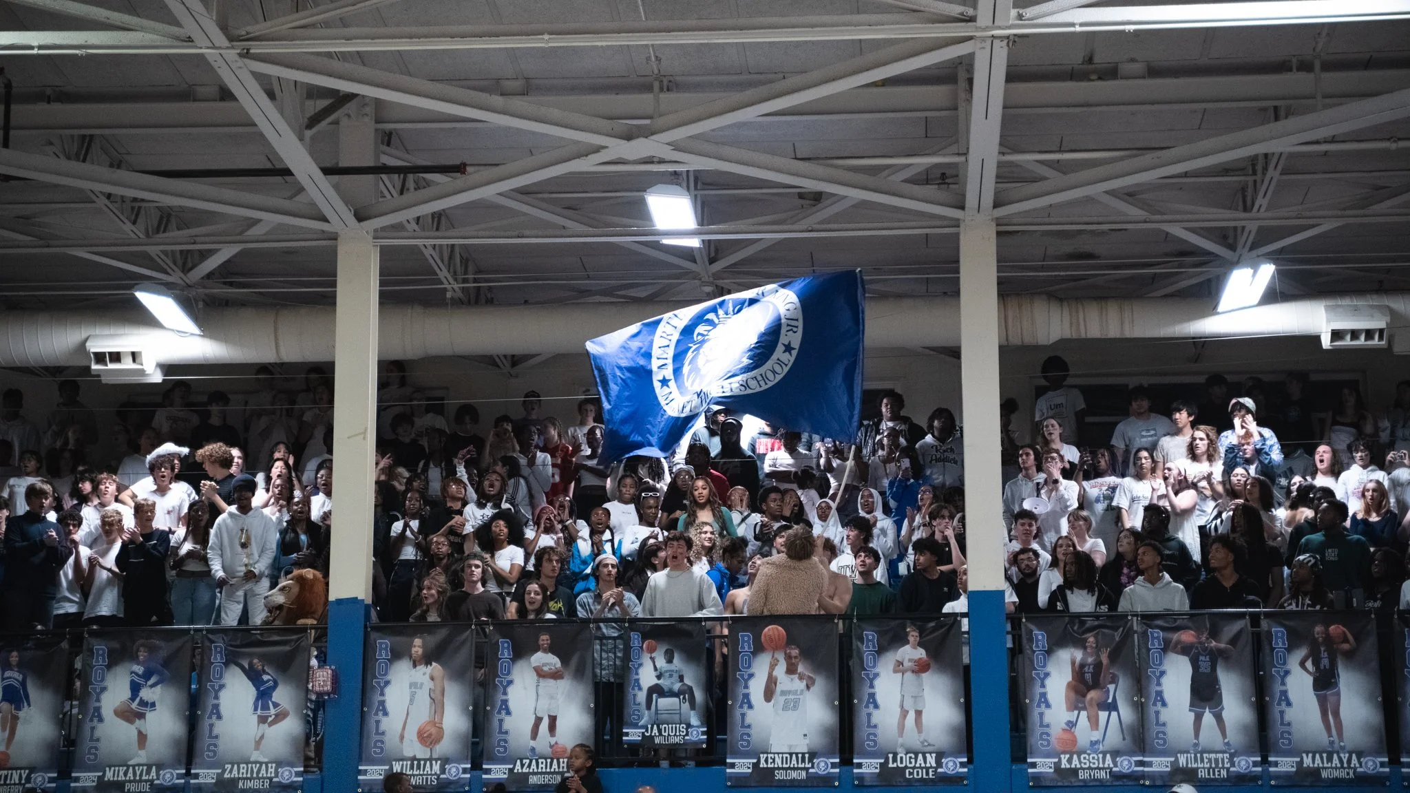 Indoor arena filled with a large crowd of students, many wearing white shirts, cheering and reacting. A blue flag with a white emblem is being waved above the crowd. The background features a high industrial ceiling with bright overhead lights.