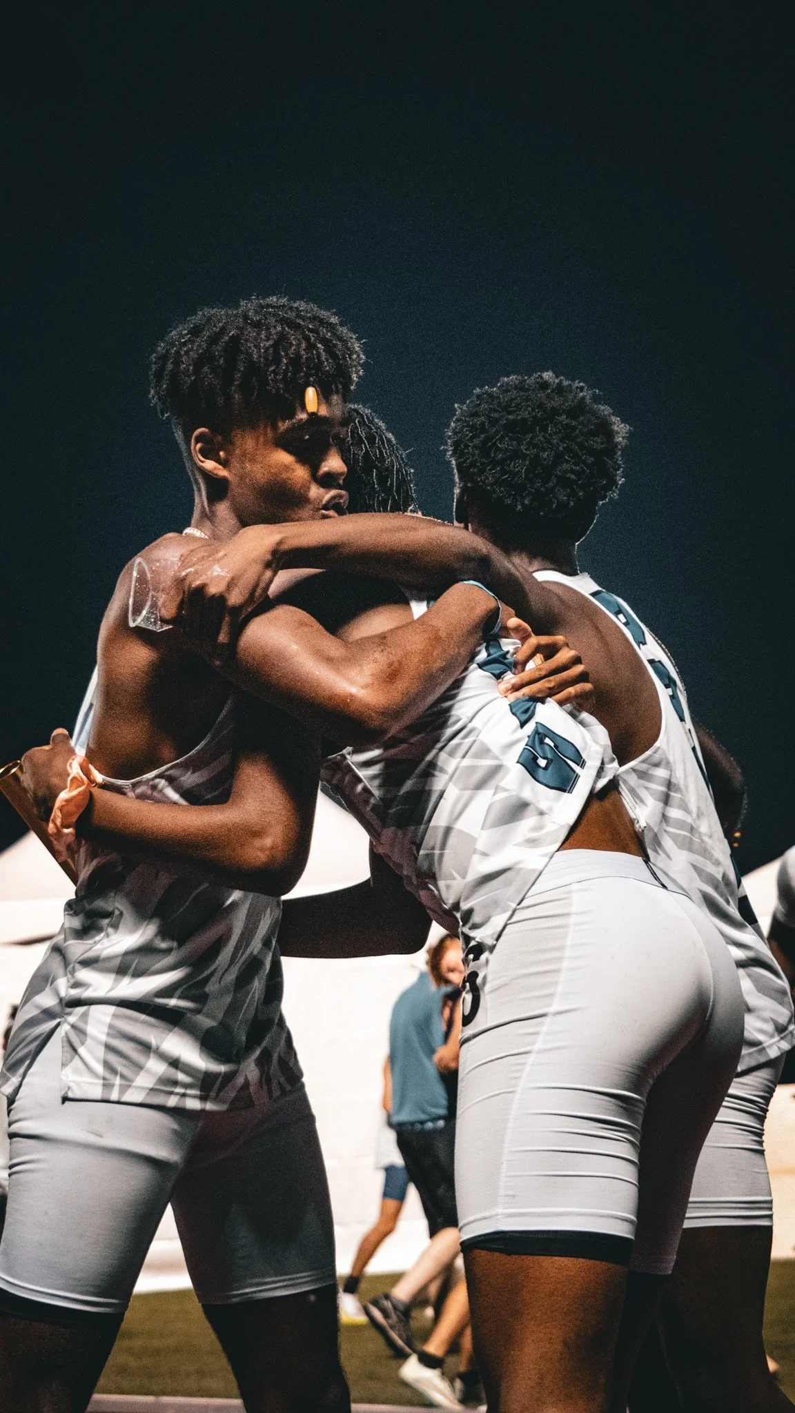 Two basketball players hugging during a game, wearing gray and white uniforms, in a stadium under dark lights.