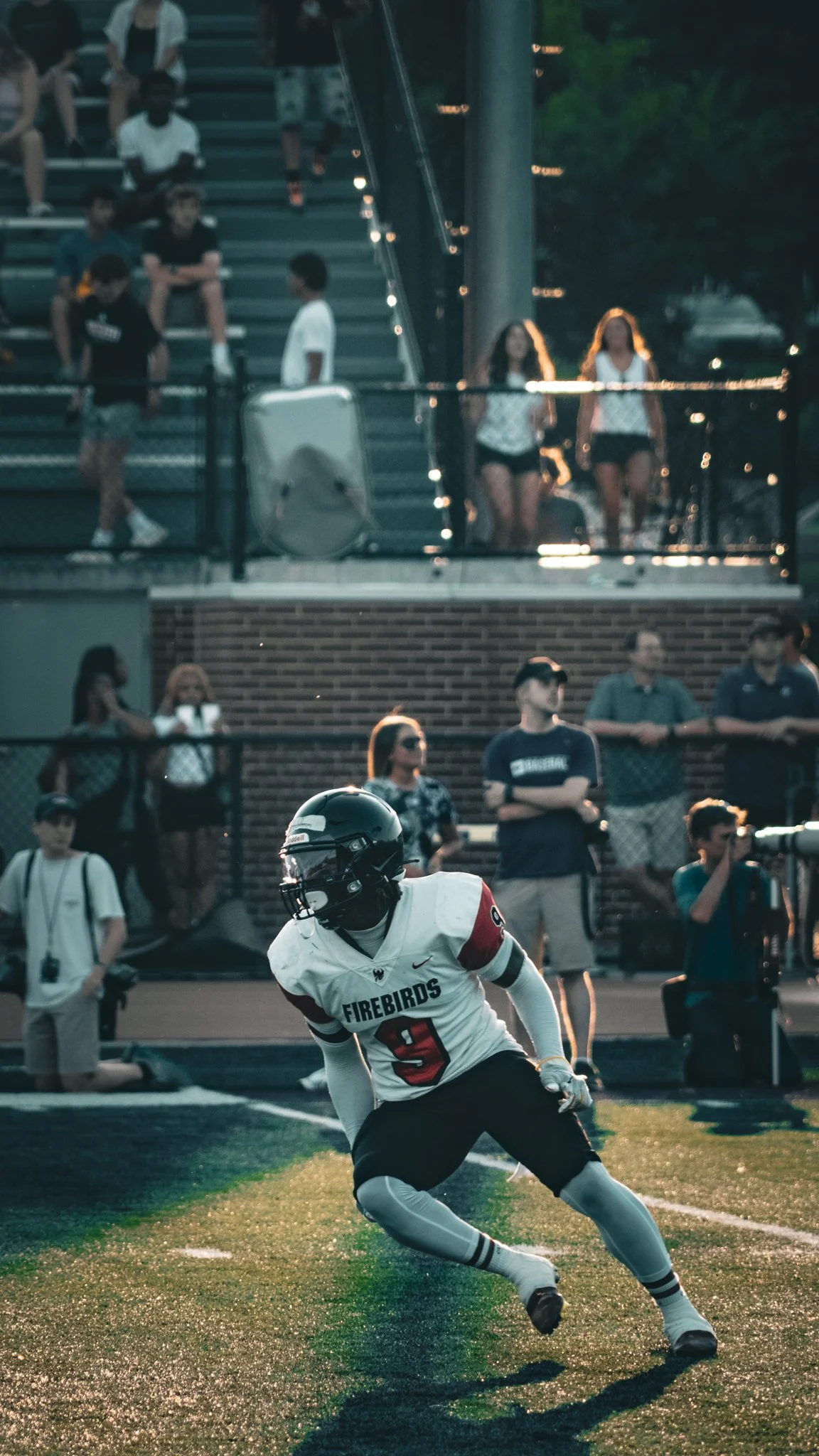 A football player in a white jersey with the word "Firebirds" and the number 9 is on the field in a game or practice, with spectators and other individuals watching from the bleachers and sidelines during evening or late afternoon.