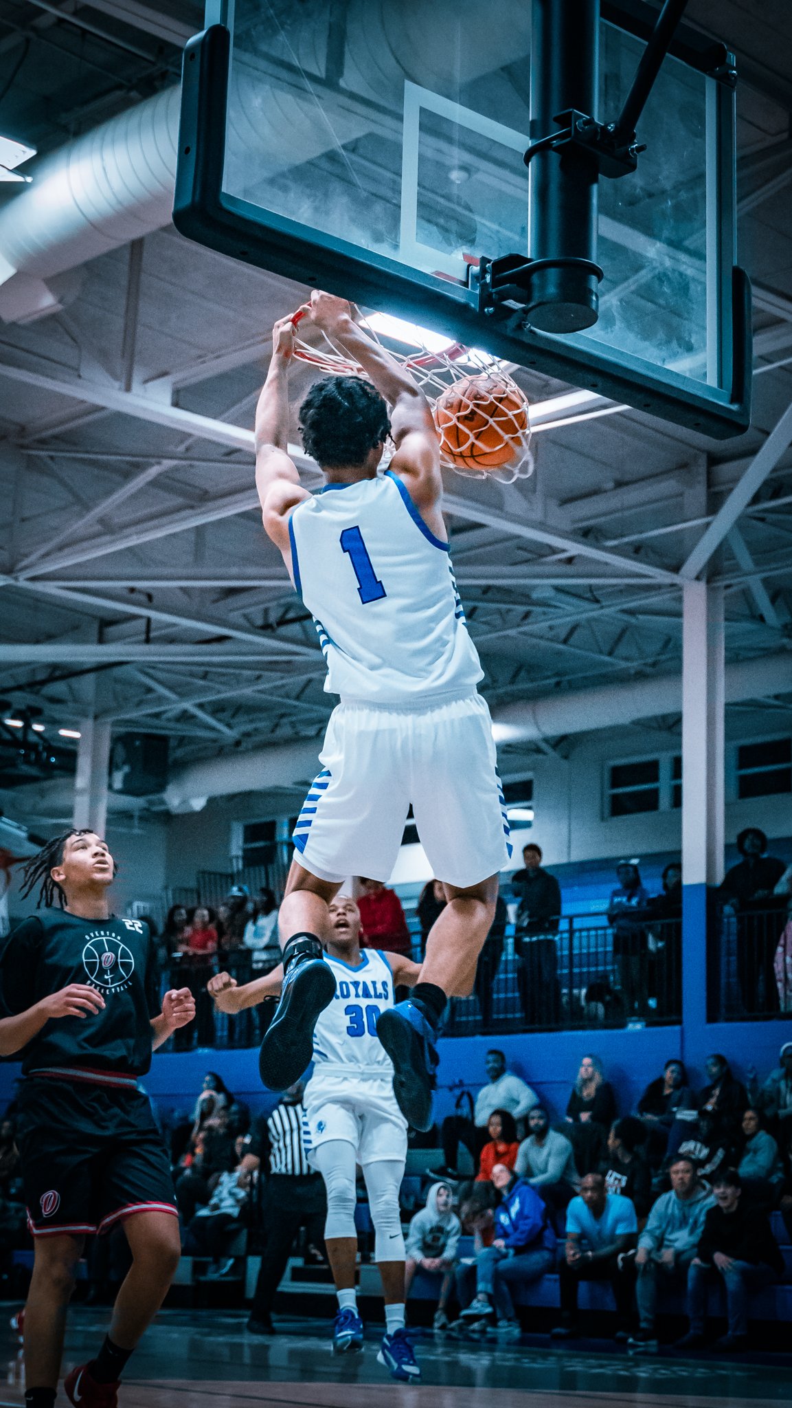 A basketball player wearing a white jersey with the number 1 is jumping to make a shot at the hoop, with a basketball in his hands. Opponents and spectators are seen around him in an indoor basketball court.